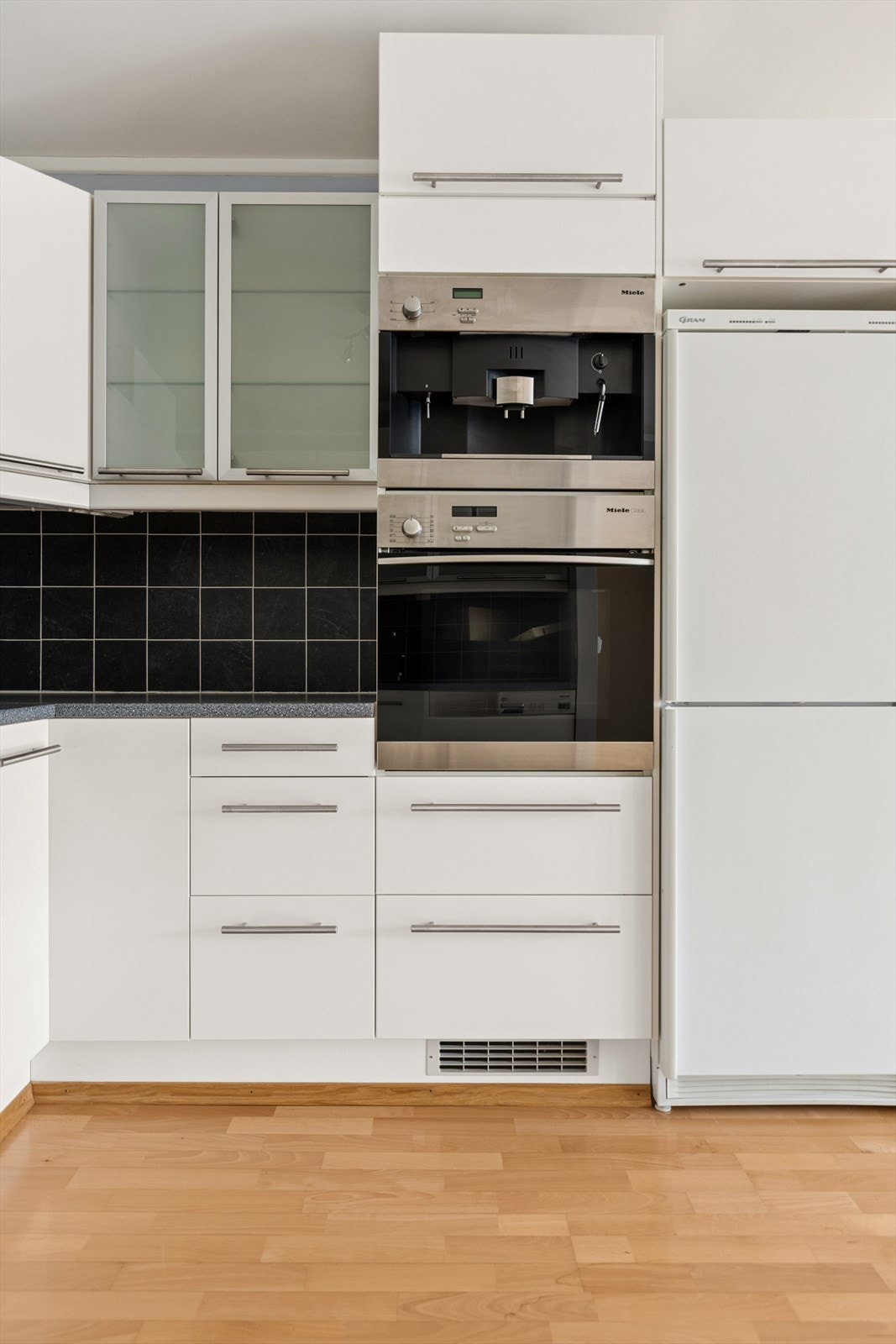 Kjøkkeninnredning med slette lakkerte fronter og enkelte glassfronter, laminat benkeplate med fliser på bakvegg. Innfelte hvitevarer bestående av stekeovn og kaffemaskin. Galleribilde
