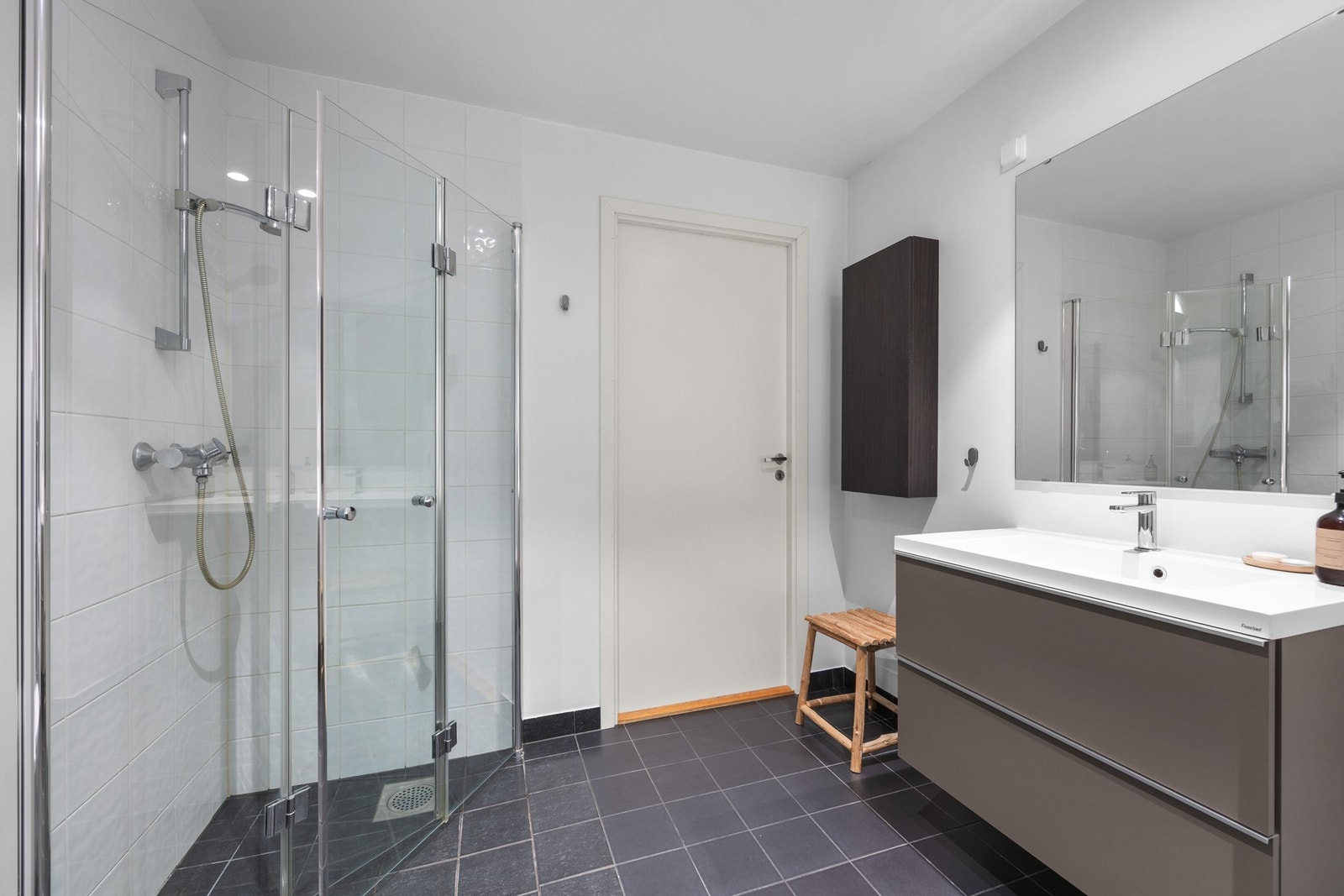 Badet har speil, heldekkende servant på møblement, vegghengt toalett og et nedsenket dusjhjørne med glassdører. Galleribilde