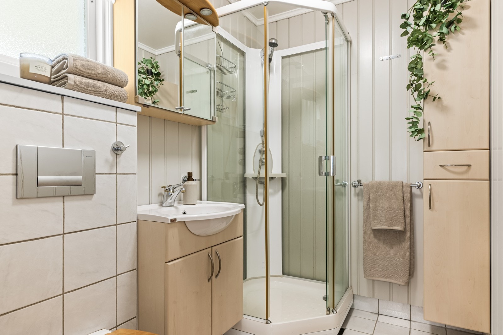 Badet i første etasje har fliser på gulv og vegger. Rommet er utstyrt med dusjkabinett og en klassisk servantinnredning med overskap med speilfrontert. I tillegg er de høyskap for ekstra lagringsplass. Galleribilde