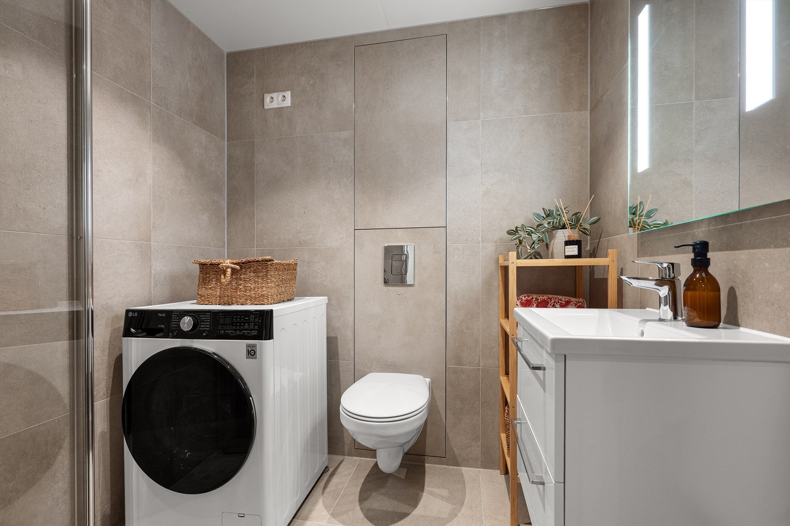 Badet er innredet med fliser på gulv og vegger, innfellbare dusjdører, vegghengt toalett, servant i skuffeinnredning, speil med belysning og opplegg for vaskemaskin. Galleribilde