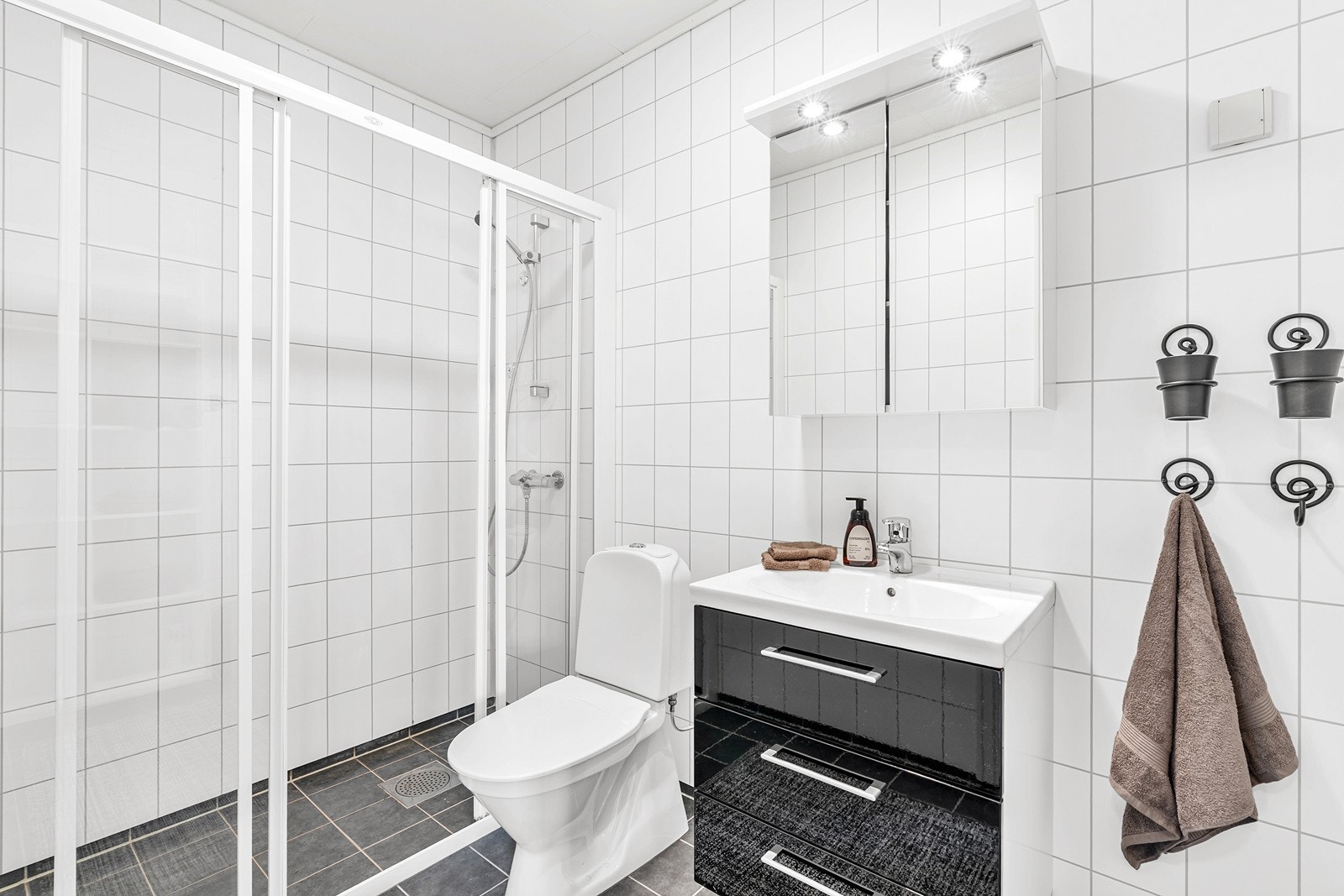 Badet har baderomsplater på vegger og fliser på gulv med elektriske varmekabler. Rommet er innredet med dusjhjørne, toalett, speilskap og heldekkende servant med skuffeseksjon. Galleribilde