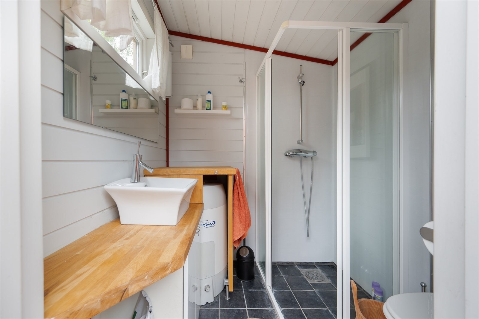 Badet inneholder et dusjhjørne med dusjkant og vegger, et snurretoalett, et gulvstående baderomsmøbel med vask, samt en varmtvannsbereder . Galleribilde