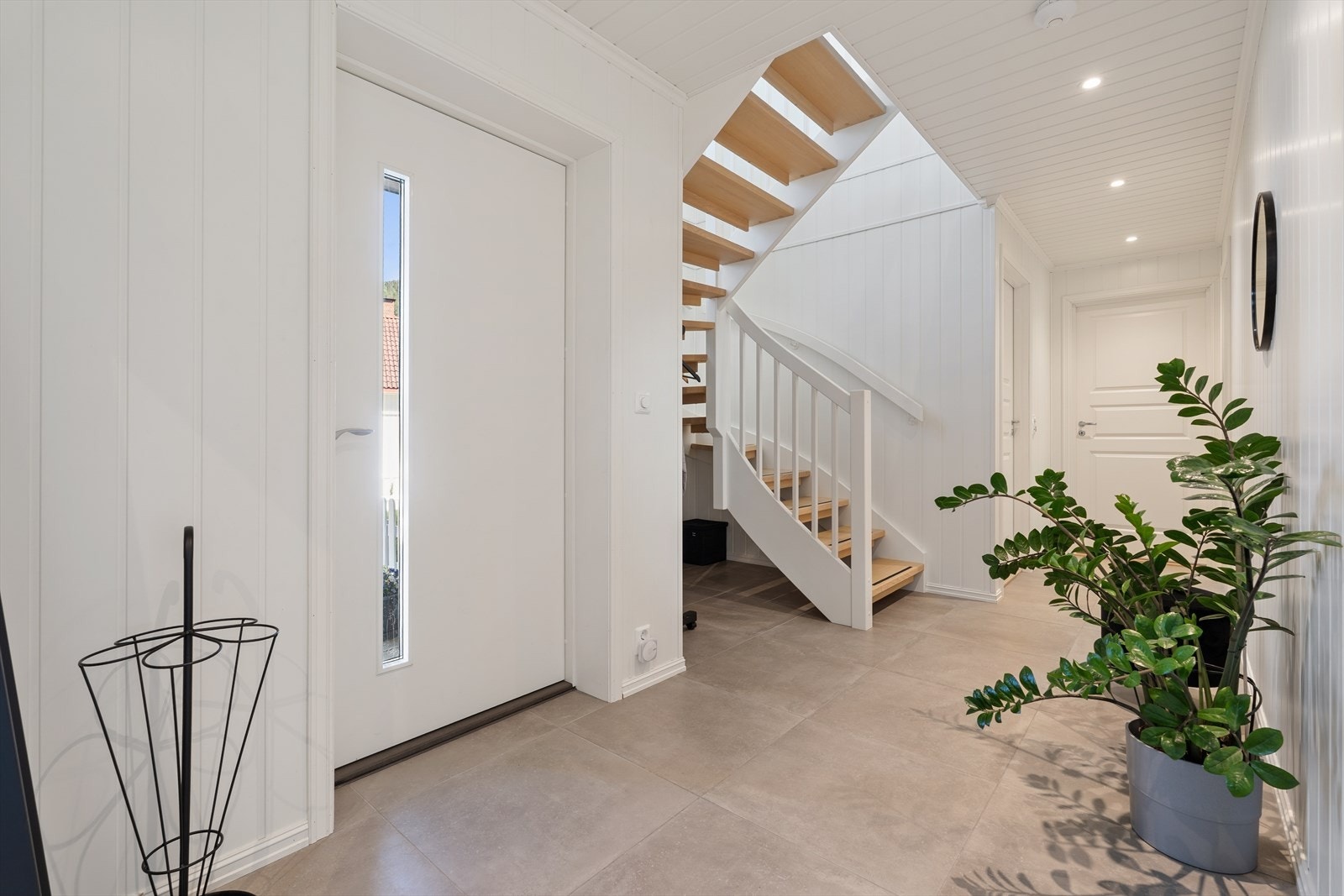 Hallen har flislagt gulv med elektriske varmekabler og en malt tretrapp som leder til andre etasje. Galleribilde