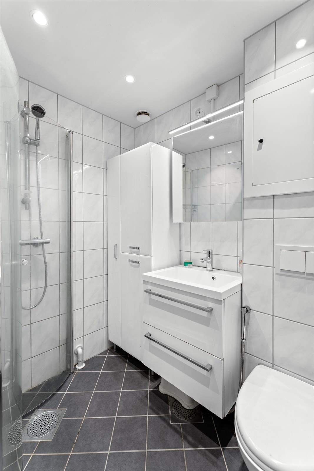 Badet er innredet med servantinnredning, veggmontert toalett og dusjhjørne med innfellbare dusjvegger i herdet glass. Speilskap med belysning gir praktisk oppbevaringsplass. Bad er rehabilitert i regi av borettslaget i 2018. Galleribilde
