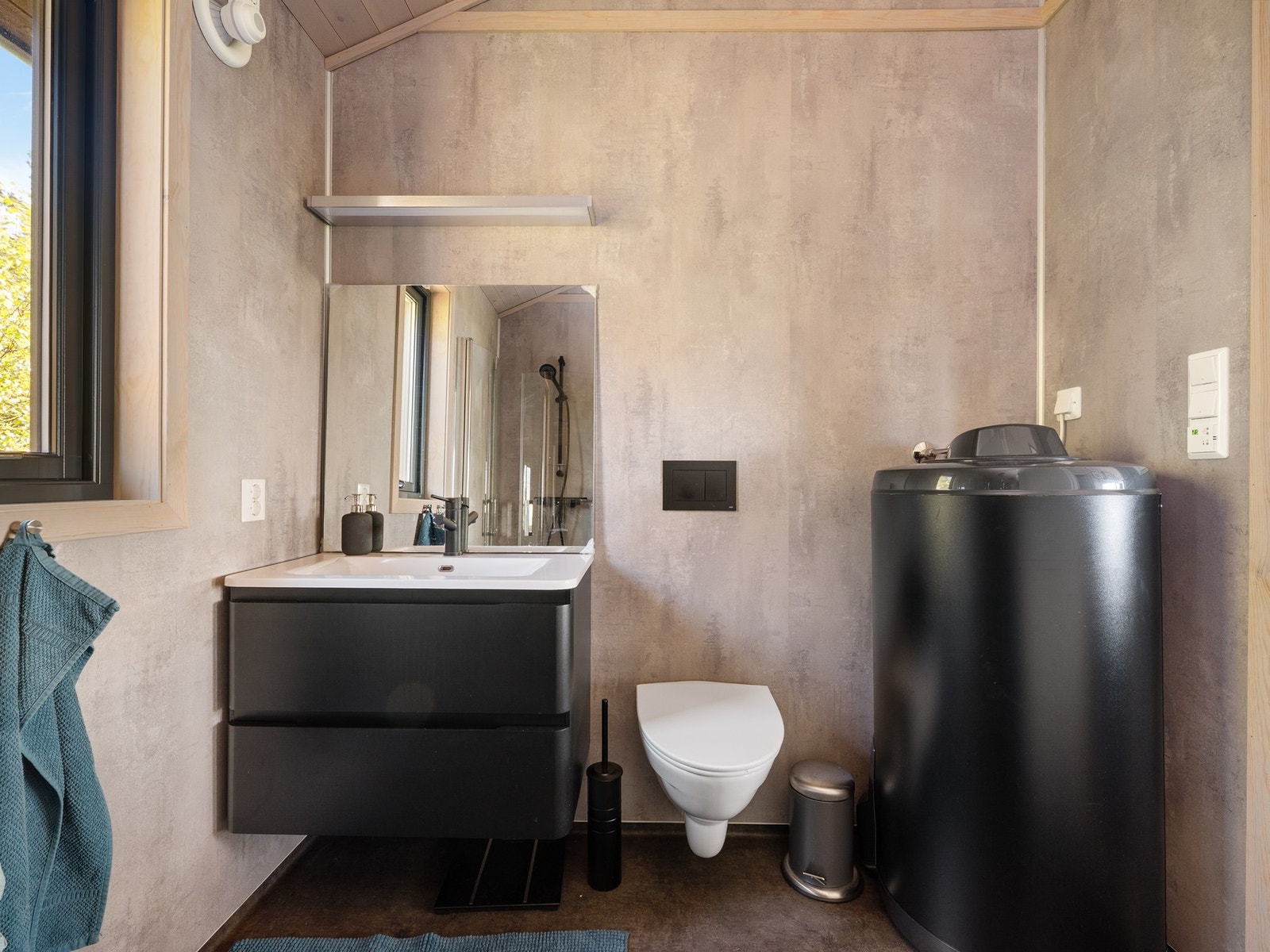 Rommet har elektriske varmekabler, avtrekksvifte og overflater med baderomsplater og vinylbelegg. Badet er innredet i lune gråtoner med sorte detaljer Galleribilde