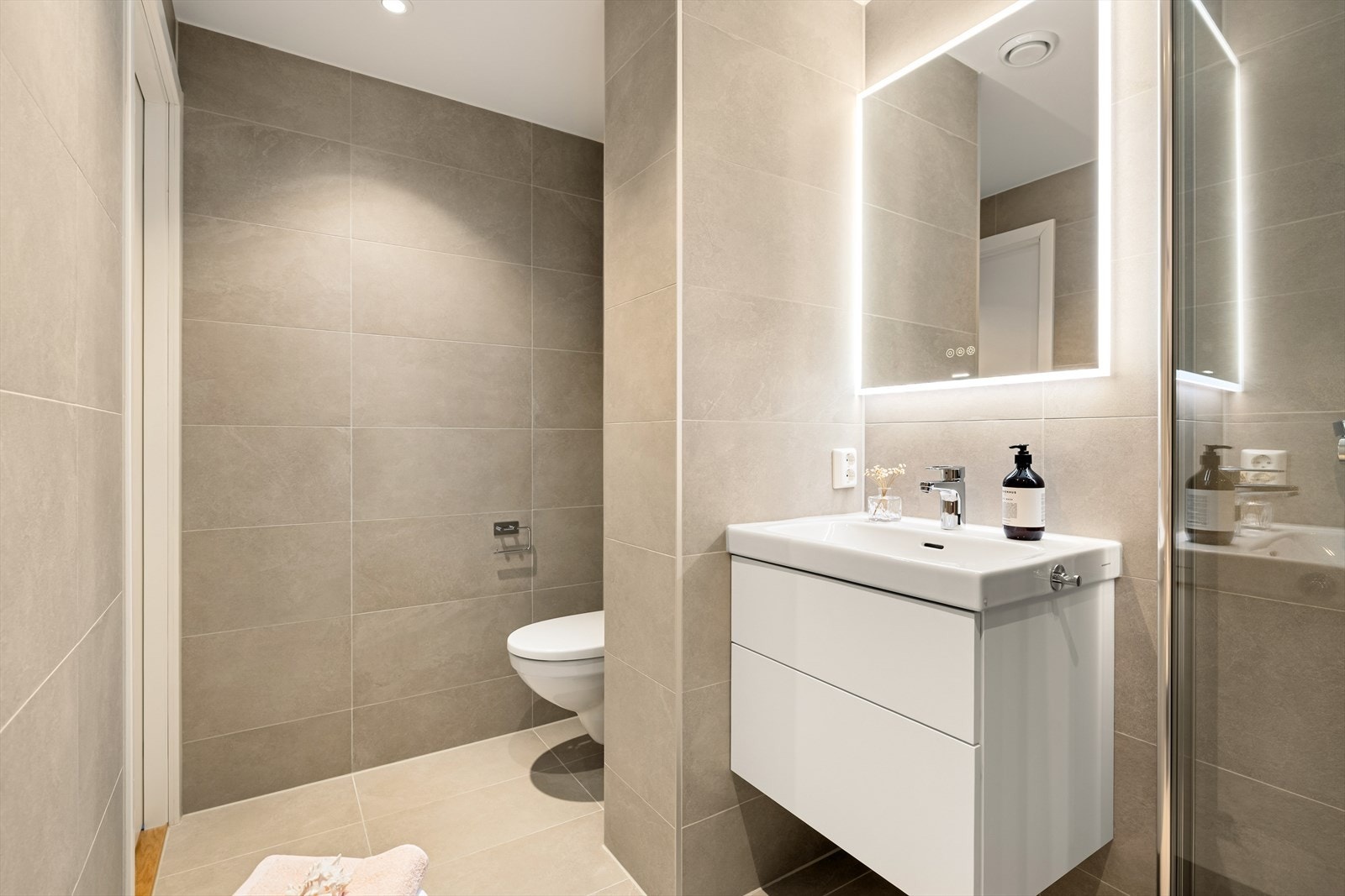 Badet inneholder baderomsinnredning med nedfelt servant, vegghengt toalett, dusjvegger og opplegg for vaskemaskin Galleribilde