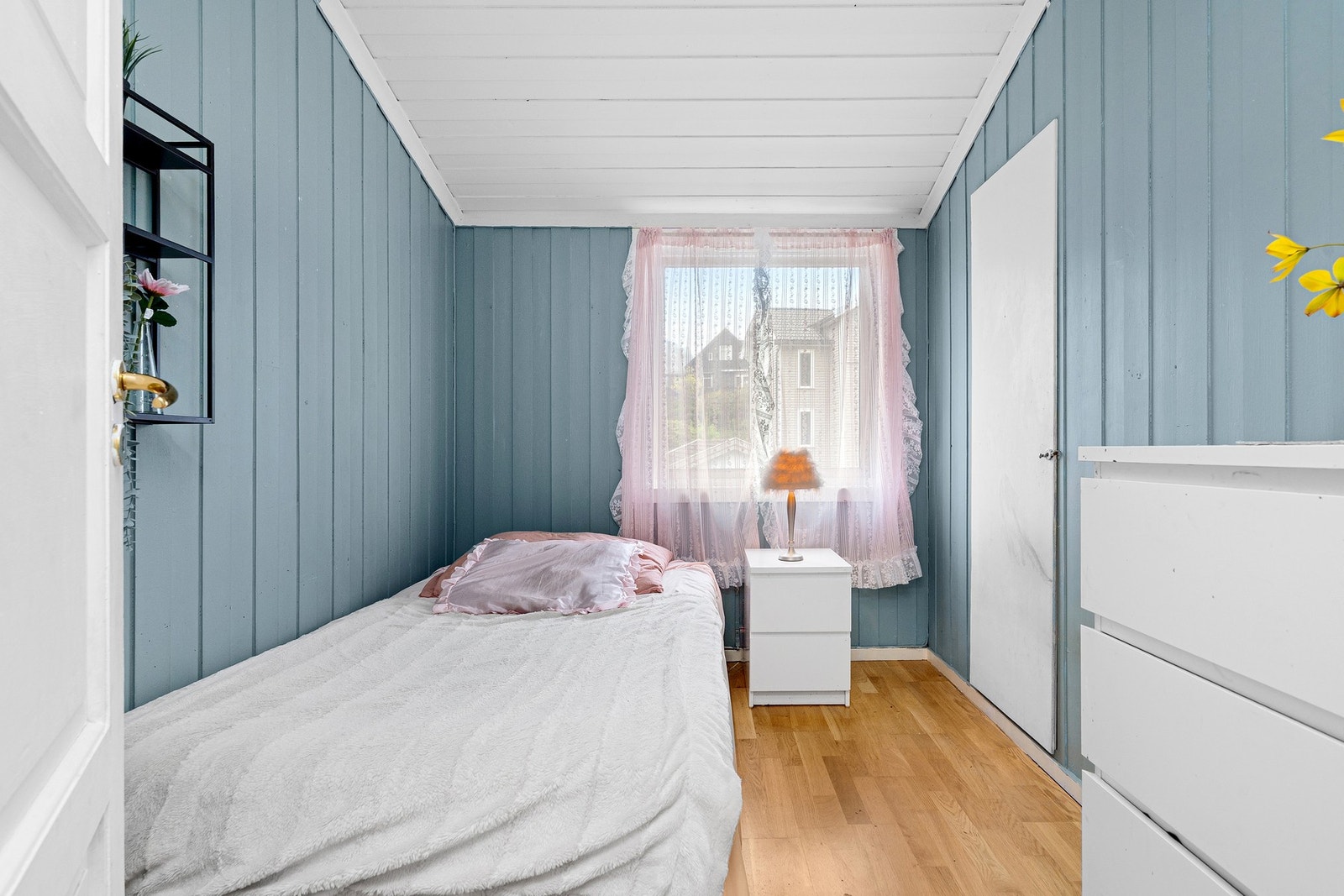 Soverom med blåmalte panelvegger, vindu med rosa gardiner og lyst tregulv. Galleribilde