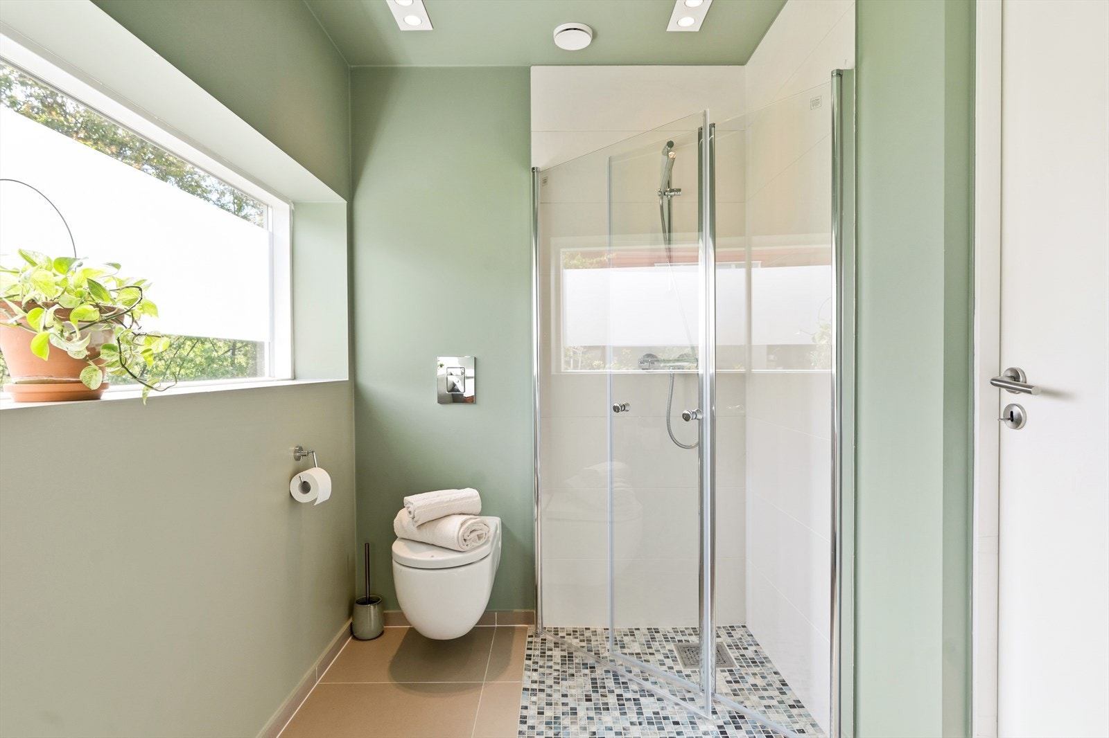 Badet har dusj med glassvegger, veggmontert toalett og lyse fliser. Rommet fremstår praktisk, pent og tidløst. Galleribilde