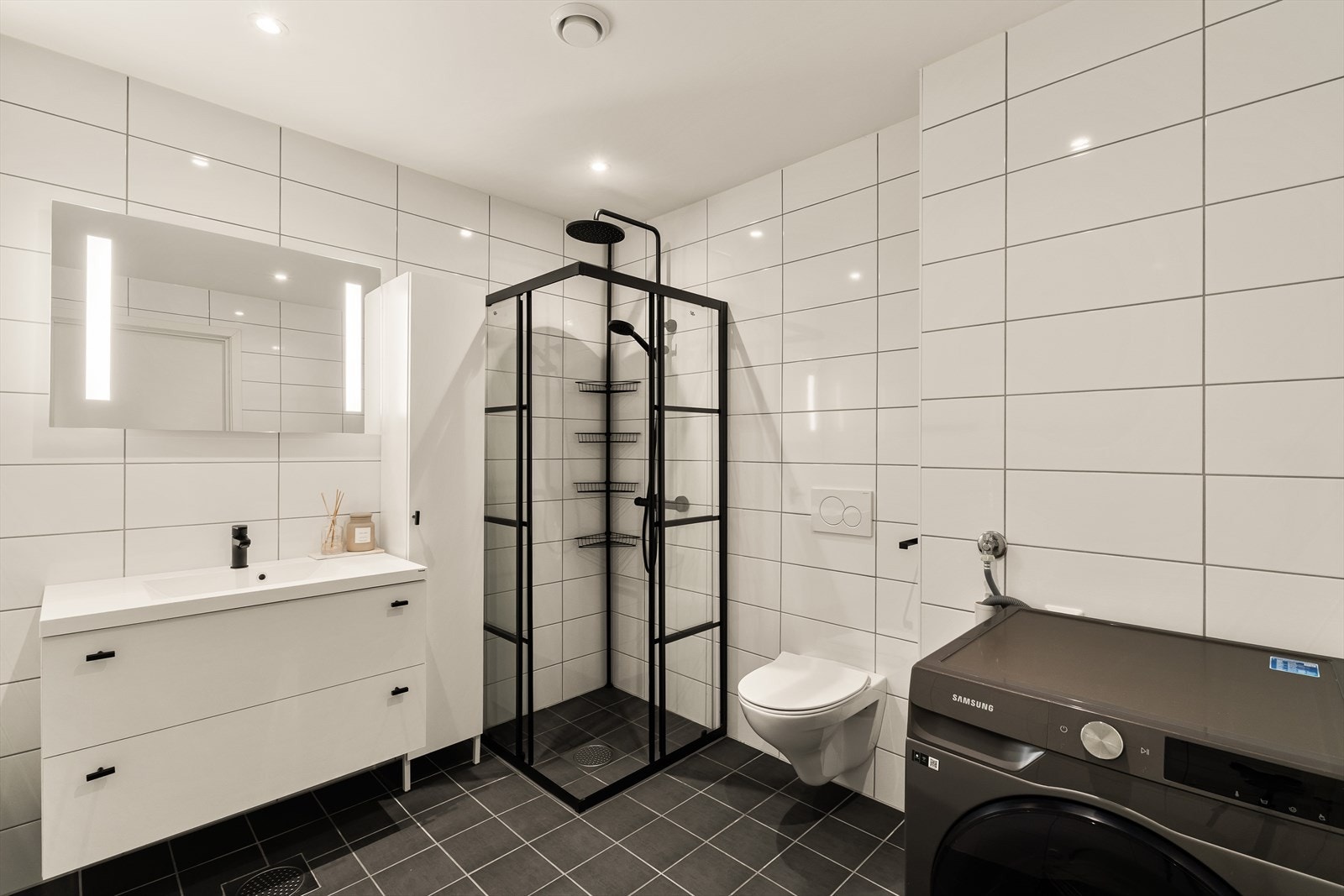 Badet er pent helfliset og inneholder servant med møblement/speil, belysning, vegghengt toalett, dusjhjørne og opplegg for vaskemaskin. Galleribilde