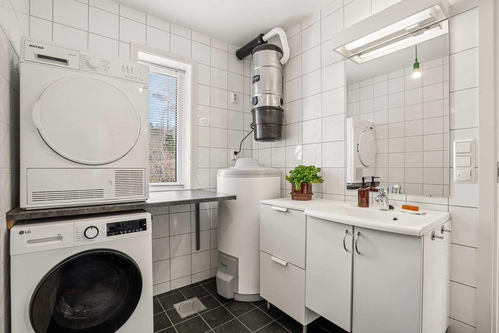 Praktisk og fint bad/vaskerom med praktisk benkeseksjon for plass til vaskemaskin og tørketrommel. På veggen ser du sentralstøvsugeren, som har uttak i begge etasjer. Galleribilde