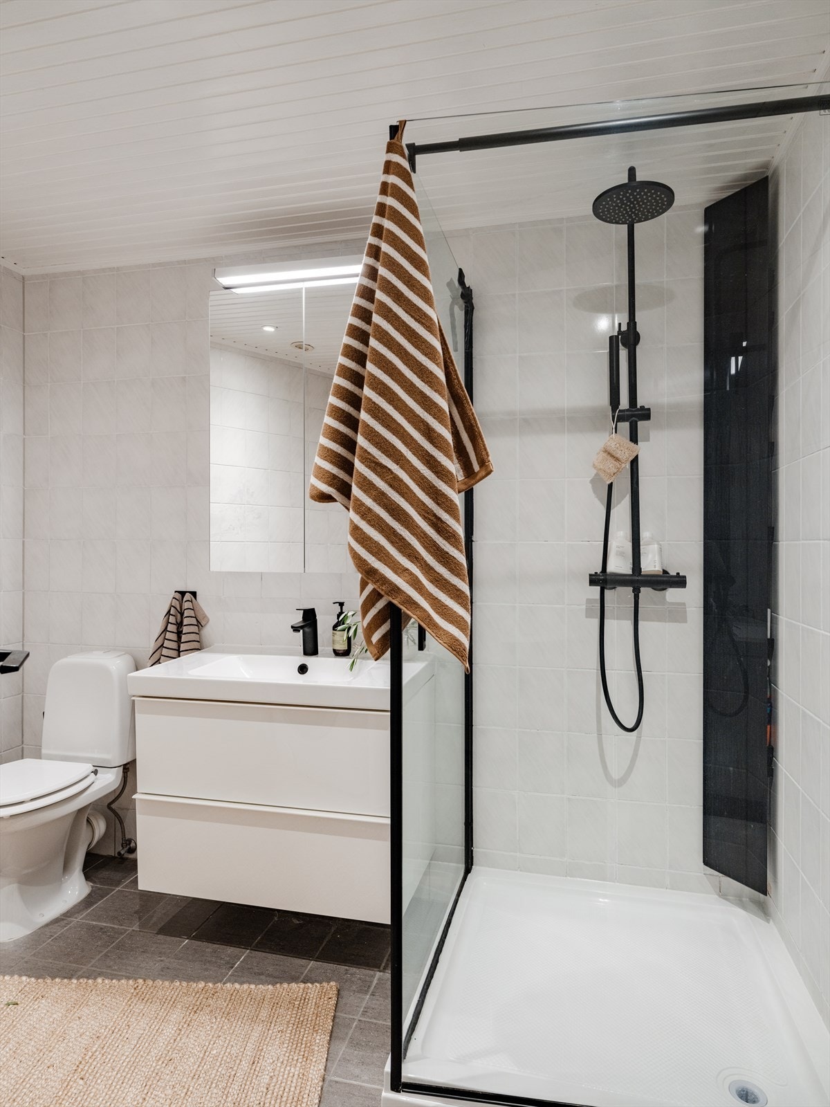 Badet er utstyrt med gulvstående toalett, servant nedfelt i innredning med oppbevaringsplass samt speilskap med belysning. Dusjkabinett med sorte detaljer og sort armatur med regnfallsdusj. Galleribilde