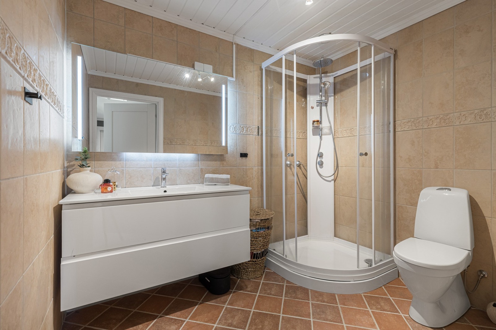 Badet fremstår pent og funksjonelt med flislagte overflater, moderne servantinnredning, speil med belysning og dusjkabinett. Galleribilde
