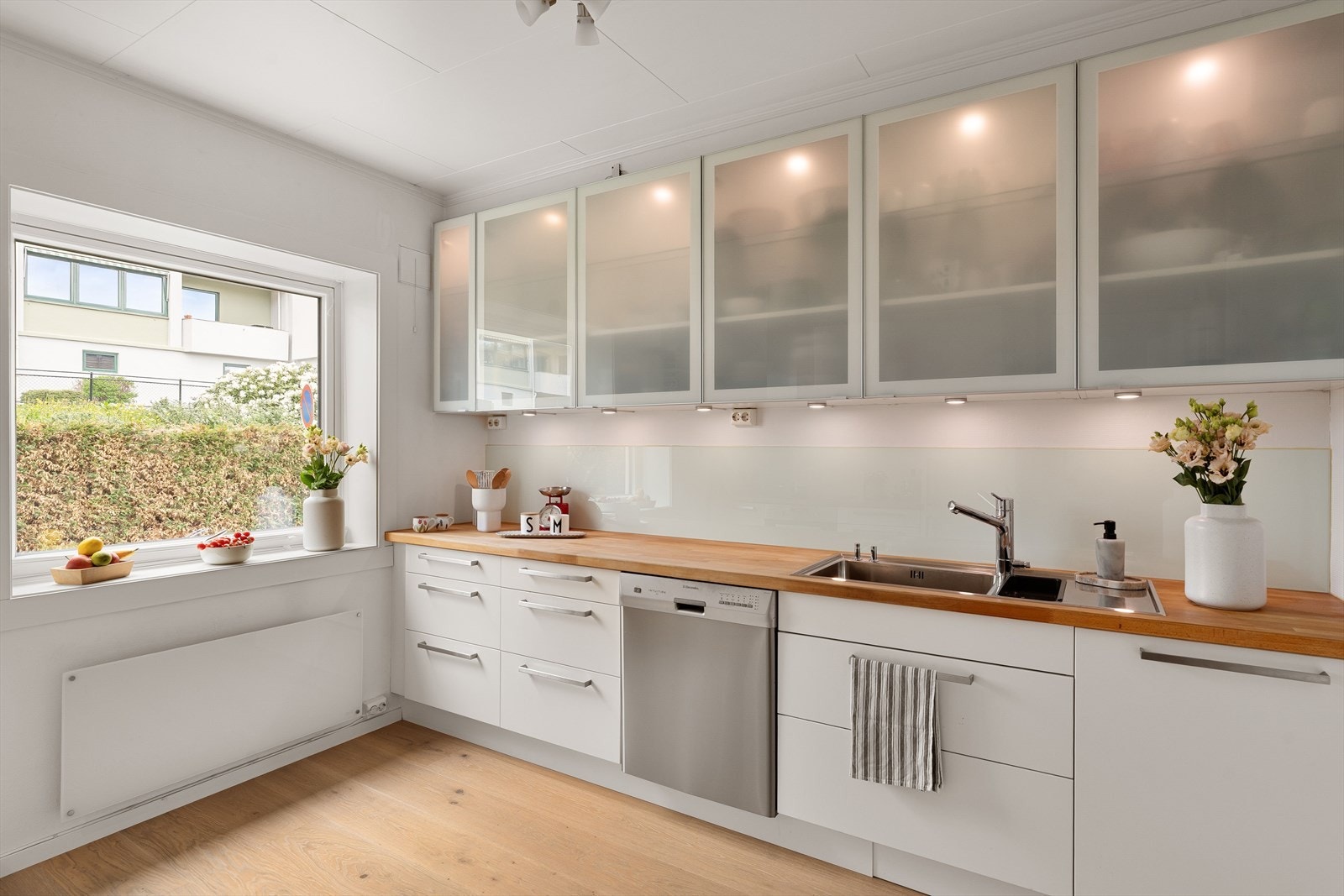 Kjøkkenet måler 10,2 kvm og er innredet med IKEA kjøkkeninnredning (2012/13) med slette hvite fronter (hvorav seks overskap i frostet glass), heltre benkeplate og glass over benkeplate. Galleribilde