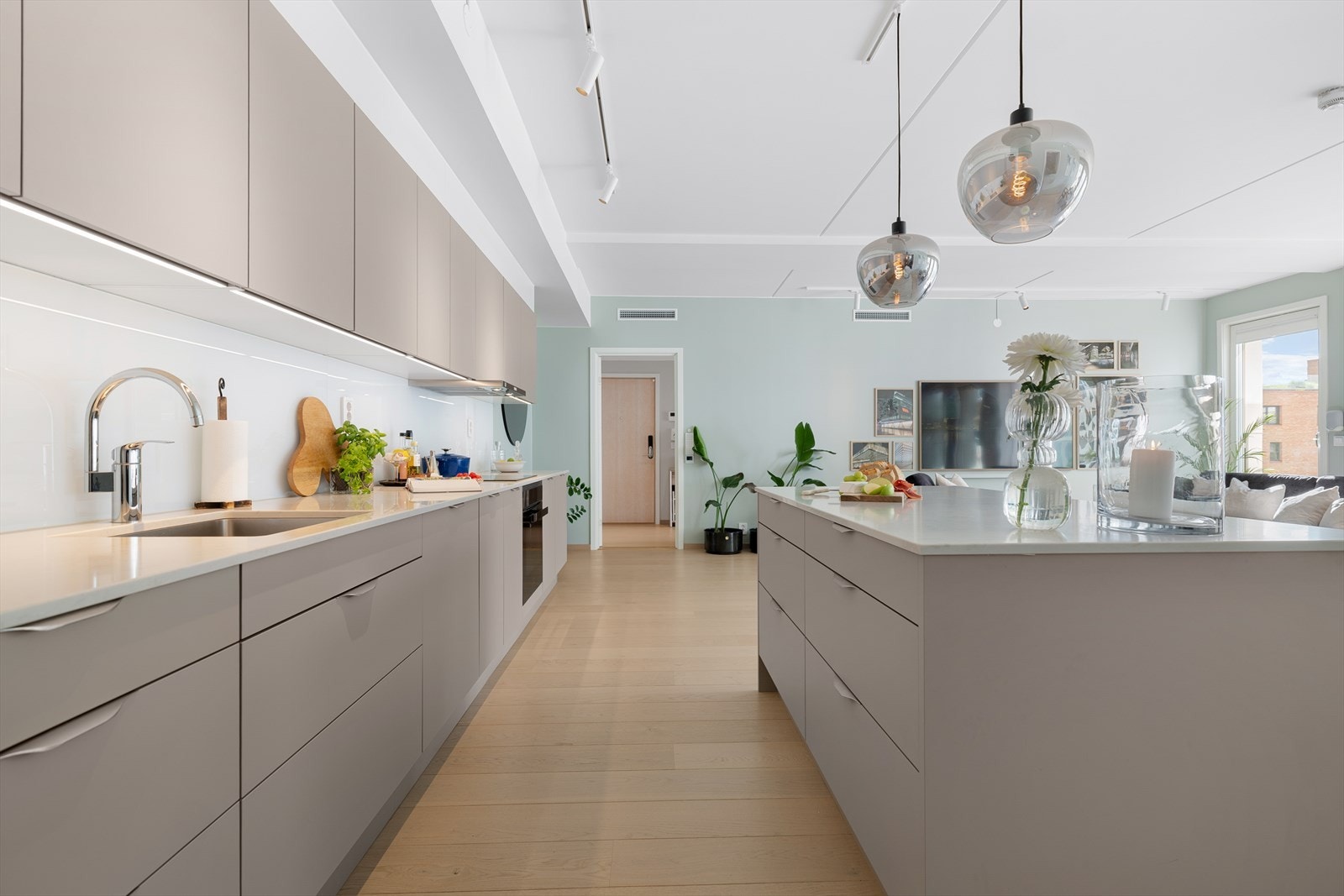 Kjøkkeninnredningen fra HTH har en dempet og harmonisk beige fargetone med hvite benkeplater i kompositt, underlimt oppvaskkum, ventilator og integrerte hvitevarer fra Siemens. Plater i glass mellom over benkeskap. Galleribilde