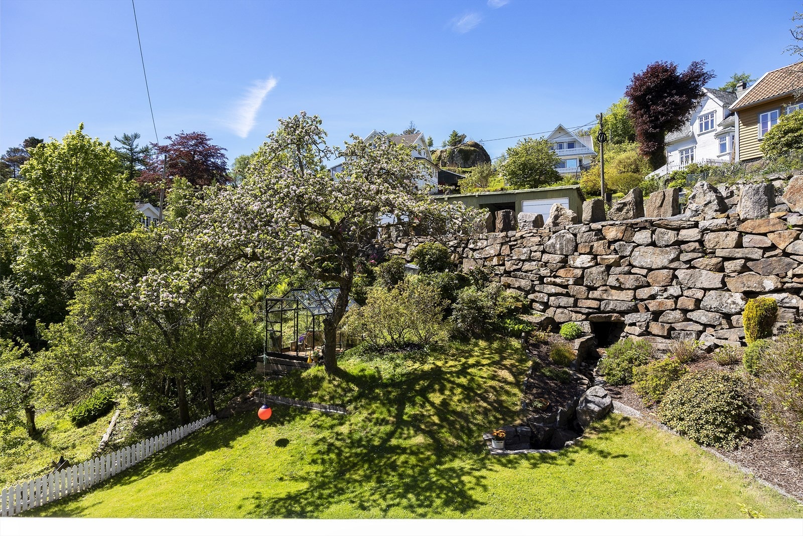 Tomten er pent opparbeidet med plen og beplantning, rammet inn av en solid natursteinsmur. Galleribilde