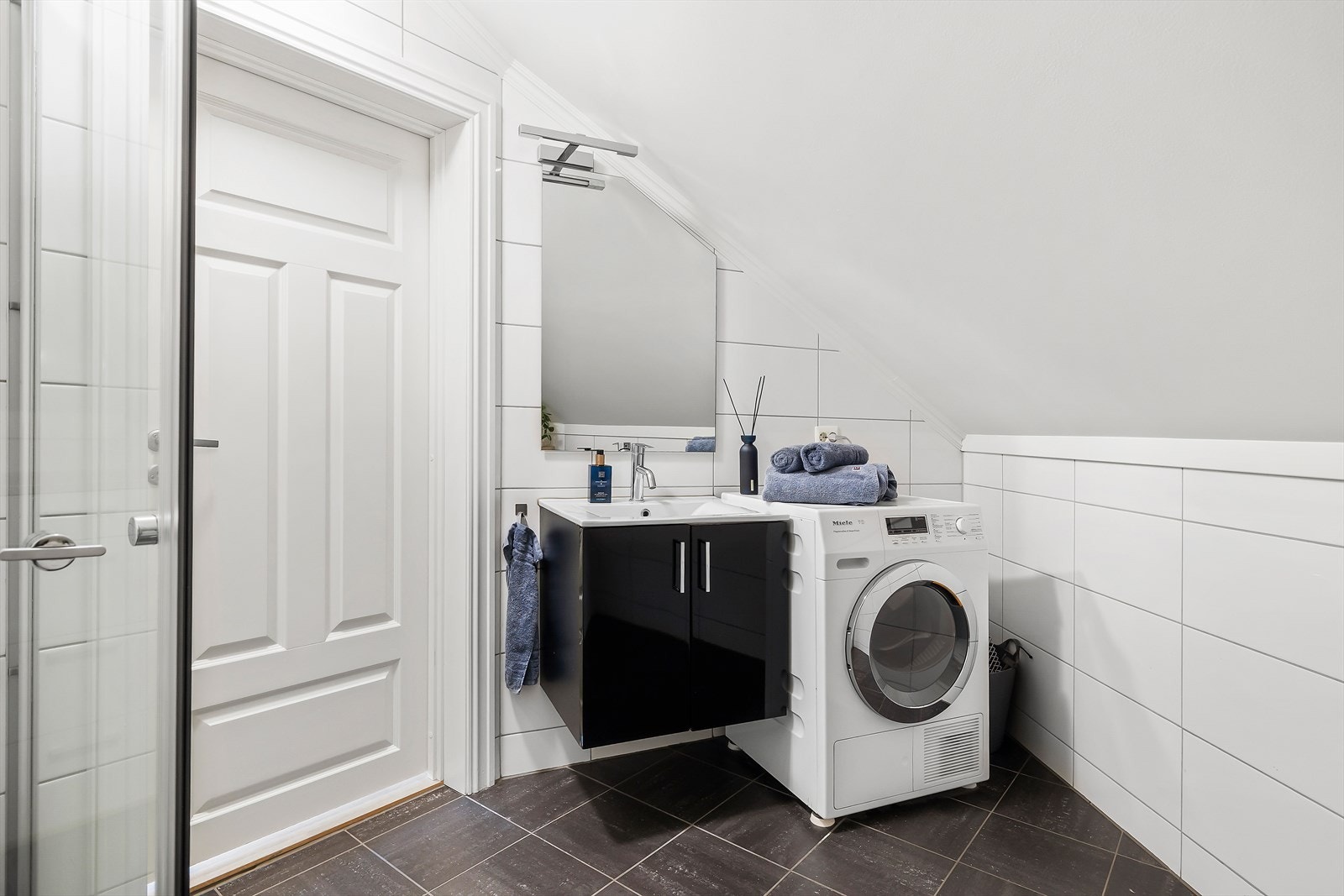 Bad i hybel fra 2012 med flis på vegg og gulv, med elektriske varmekabler og opplegg for vaskemaskin. Inneholder innredning med nedfelt servant, gulvmontert toalett og dusjhjørne med glassvegger. Galleribilde