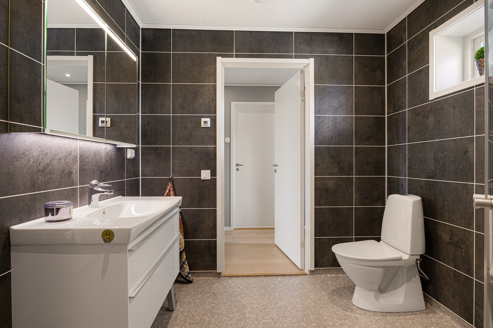 Badet er praktisk og moderne med grått gulvbelegg, mørke flisplater på veggene, gulvvarme og innfelte LED-spotter. Innredningen består av heldekkende servant på skuffemøblement, speilskap med integrert belysning og høyskap. Galleribilde
