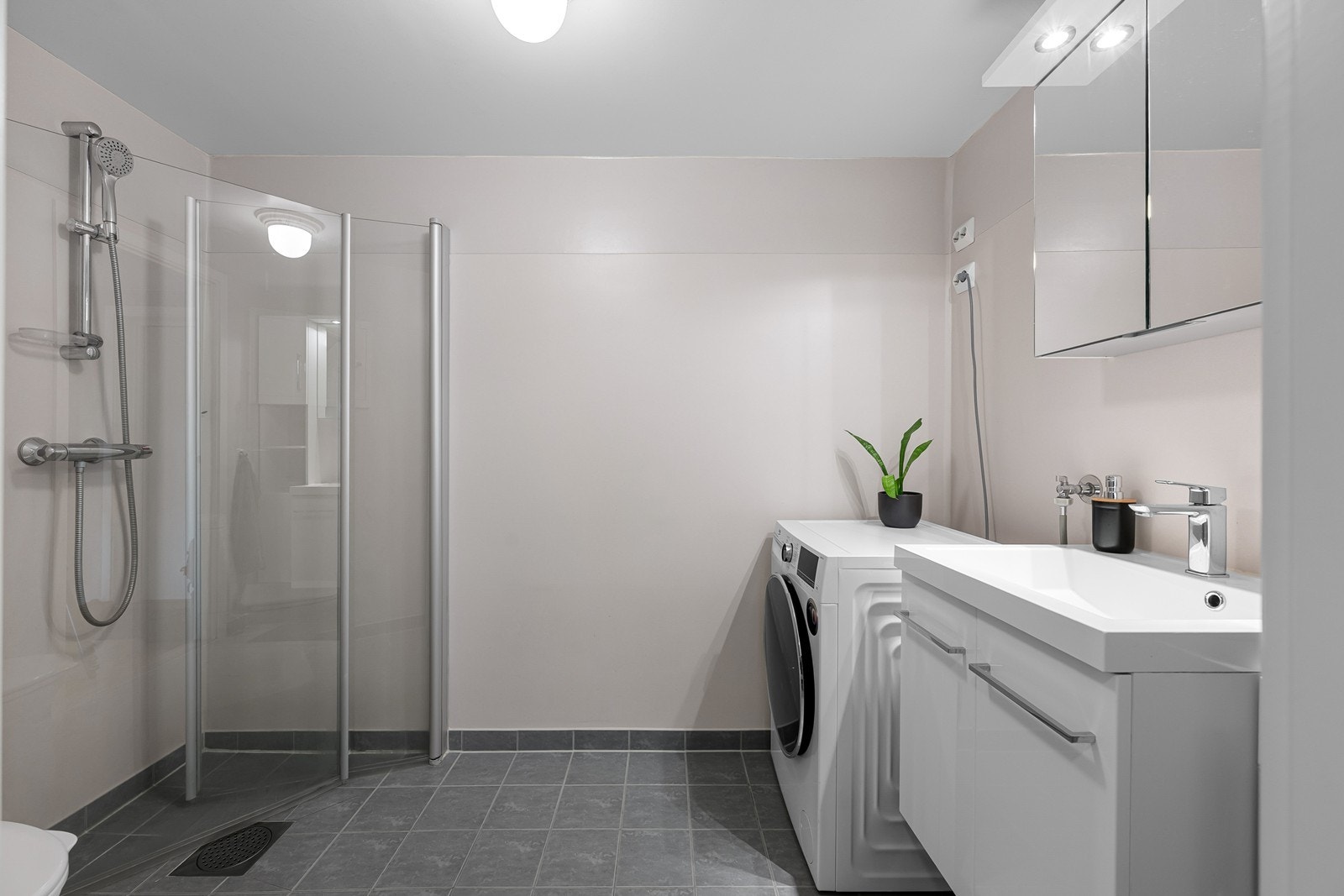 Badet er praktisk innredet med flislagt gulv, dusjløsning, servantinnredning, toalett og opplegg for vaskemaskin. Galleribilde