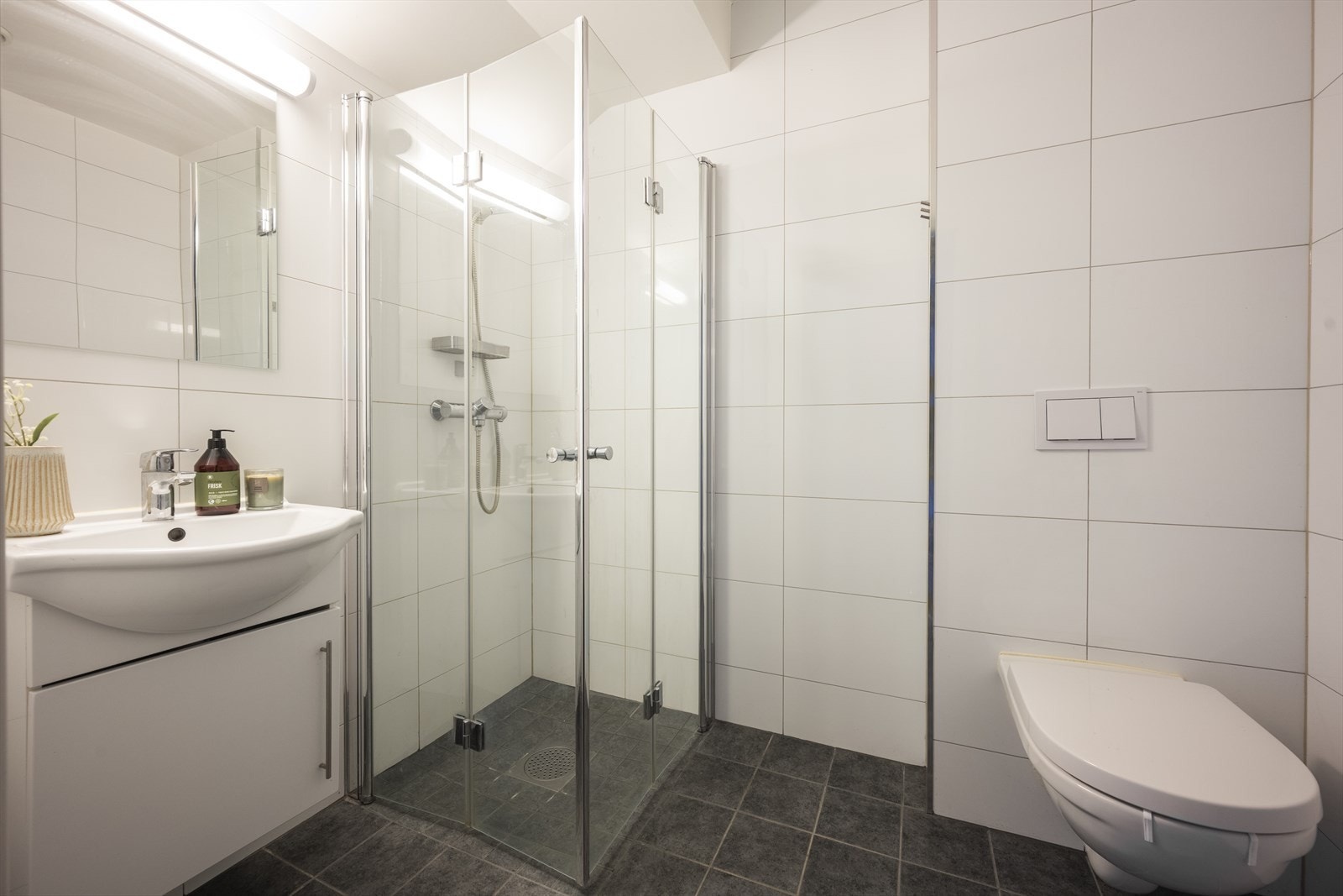 Flislagt bad med varmekabler. Badet er utstyrt med dusjhjørne med innfellbare glassdører og blandebatteri, TECE vegghengt toalett og baderomsinnredning med heldekkende servant og ettgreps blandebatteri. Galleribilde