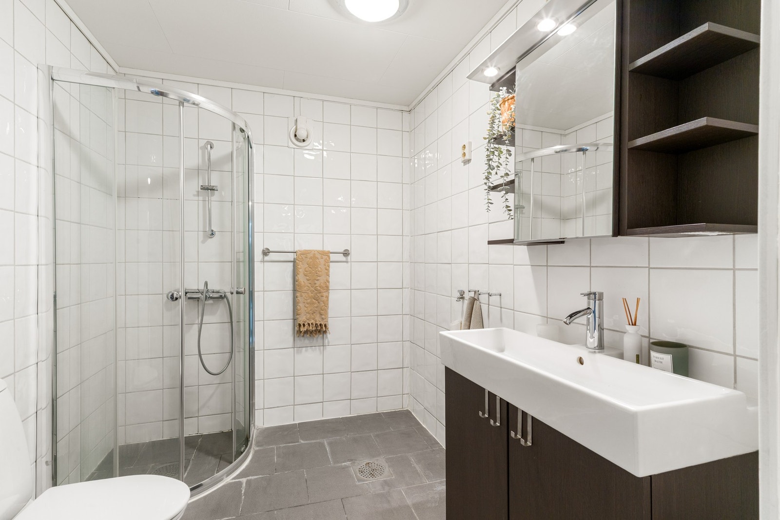 Badet har flislagte vegger og gulv med elektriske varme. Innredningen består av servant med oppbevaring, dusjhjørne, toalett og opplegg for vaskemaskin og tørketrommel. Galleribilde