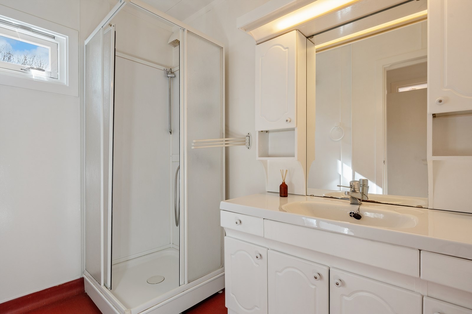 Baderommet er av eldre standard og er innredet med rødt belegg på gulv og lyse baderomsplater på vegg. Badet er ellers utstyrt med dusjkabinett og baderomsinnredning med god lagringsplass og nedfelt servant. Galleribilde