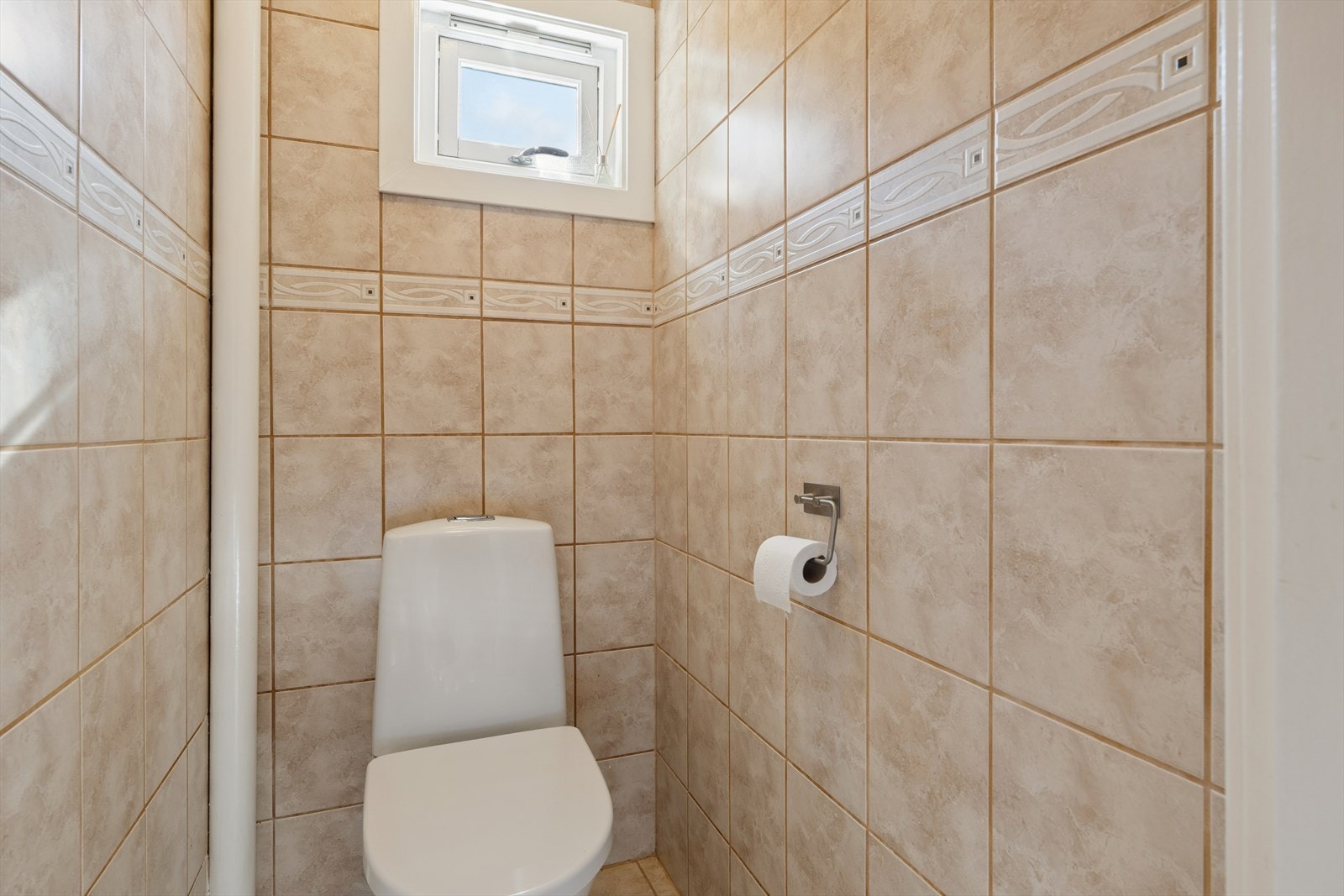 Vegg i vegg med baderommet ligger et toalettrom innredet med beige fliser på gulv og vegger. Rommet er utstyrt med et gulvmontert toalett som ble installert i 2022. Galleribilde
