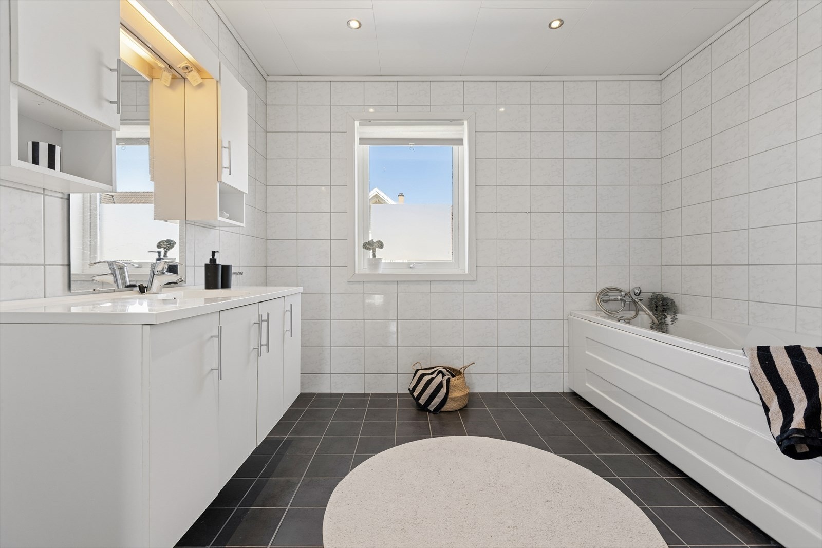 Badet har tidløse fliser på overflater i sort og hvitt. Innredningen består av servantseksjon og veggskap med hvite fronter, samt speil og ett høyskap. Galleribilde