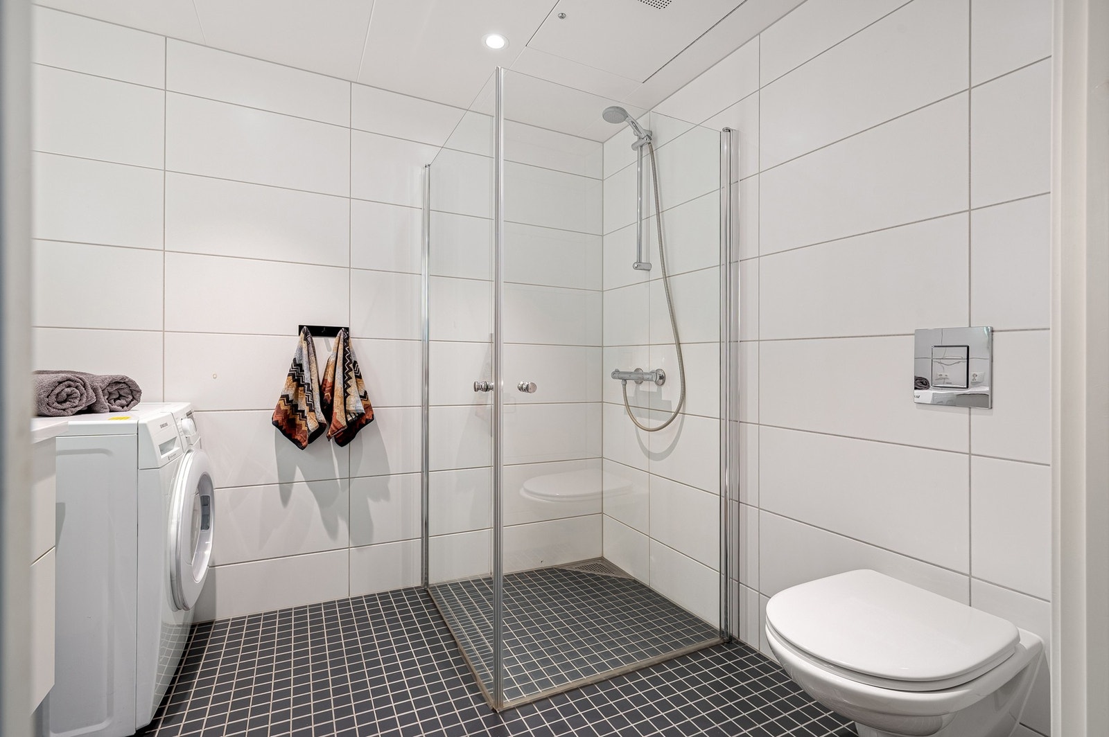 Badet fremstår lekkert og rommer gode 4,5 kvm. Innredet med plassbesparende dusjdører i glass, veggmontert wc, servantplate på underskap og speil med lys. I hjørne av badet har du kran og avløp for vaskemaskin. Galleribilde