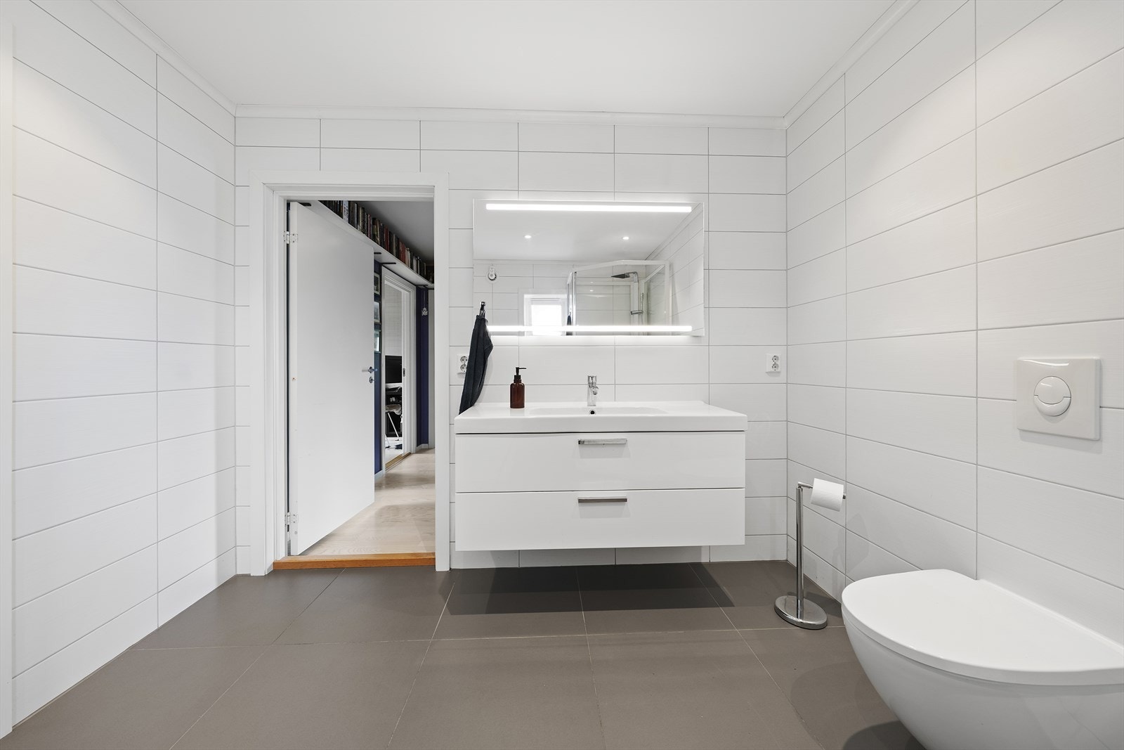 Badet er utstyrt med heldekkende servant over skuffeinnredning, speil med belysning, badekar, dusjkabinett og vegghengt toalett Galleribilde