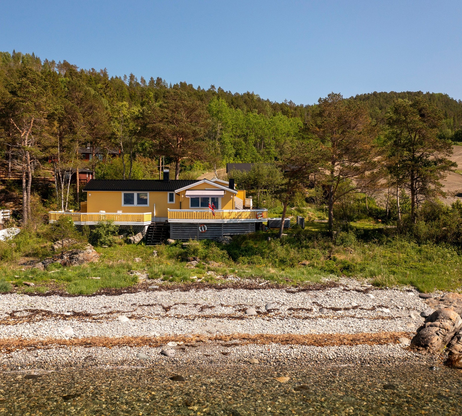 Hytta ligger tilnærmet i vannkanten og den er omkranset av skogsområder i bakgrunn. Galleribilde