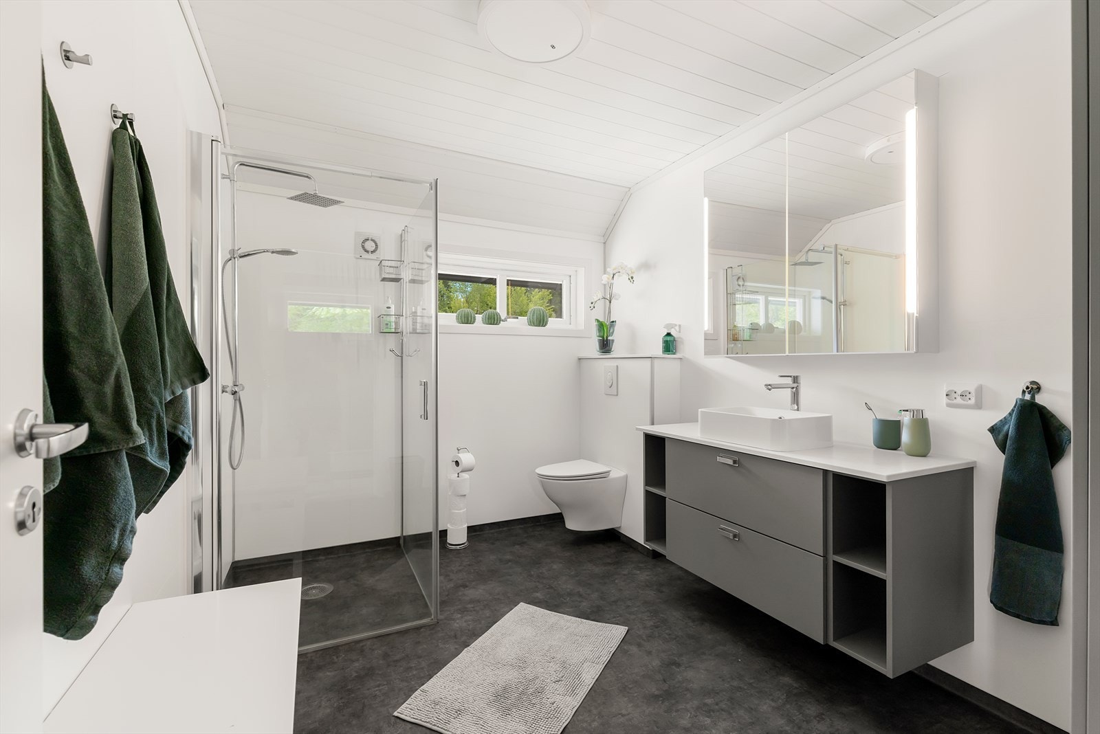 Badet fremstår stilrent med belegg på gulv og plater på veggene. Rommet innholder dusjhjørne, servant med skuffer og hyller, speilskap og vegghengt wc. Galleribilde