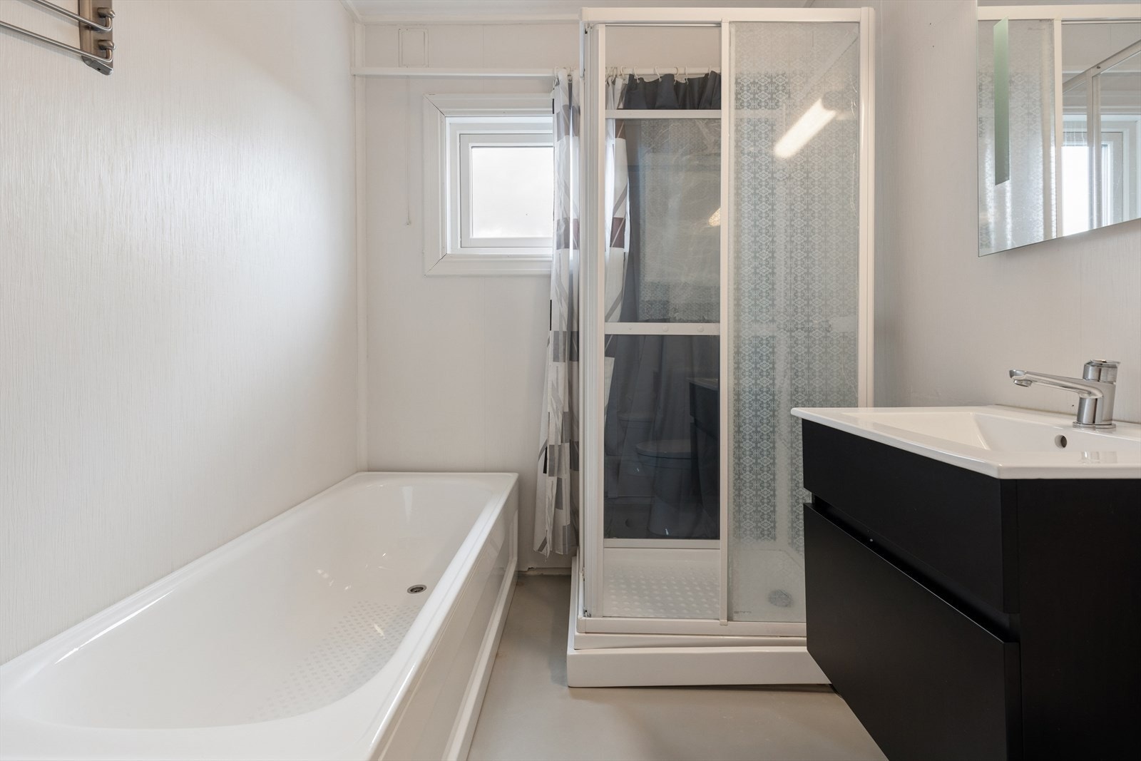 Badet har vegger tapetsert med våtromstapet og gulv har vinylbelegg. Utstyrt med gulvstående toalett, dusjkabinett, badekar og enkel servant. Galleribilde