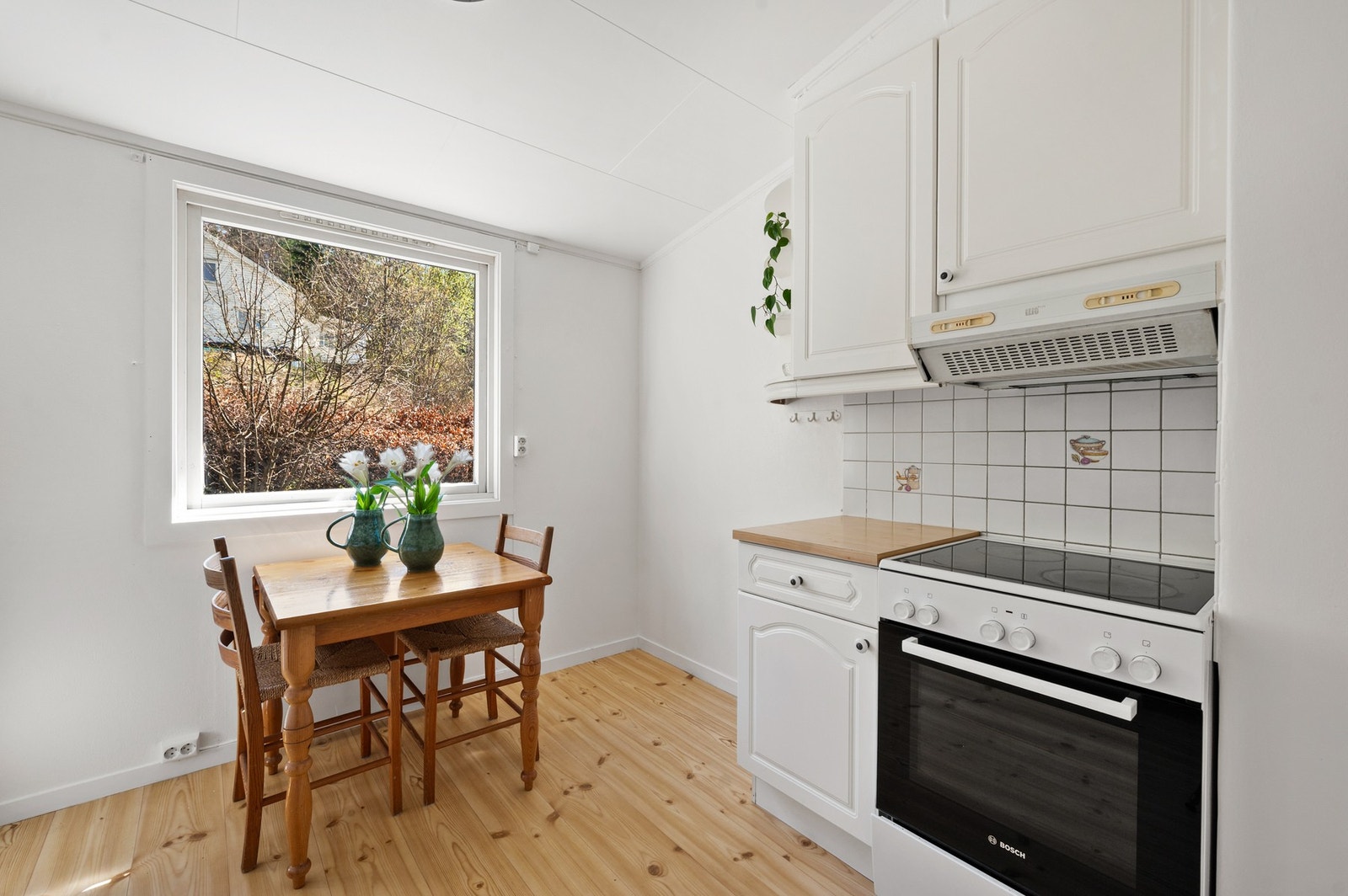 Lyst kjøkken med tregulv, hvit innredning og plass til lite spisebord. Galleribilde