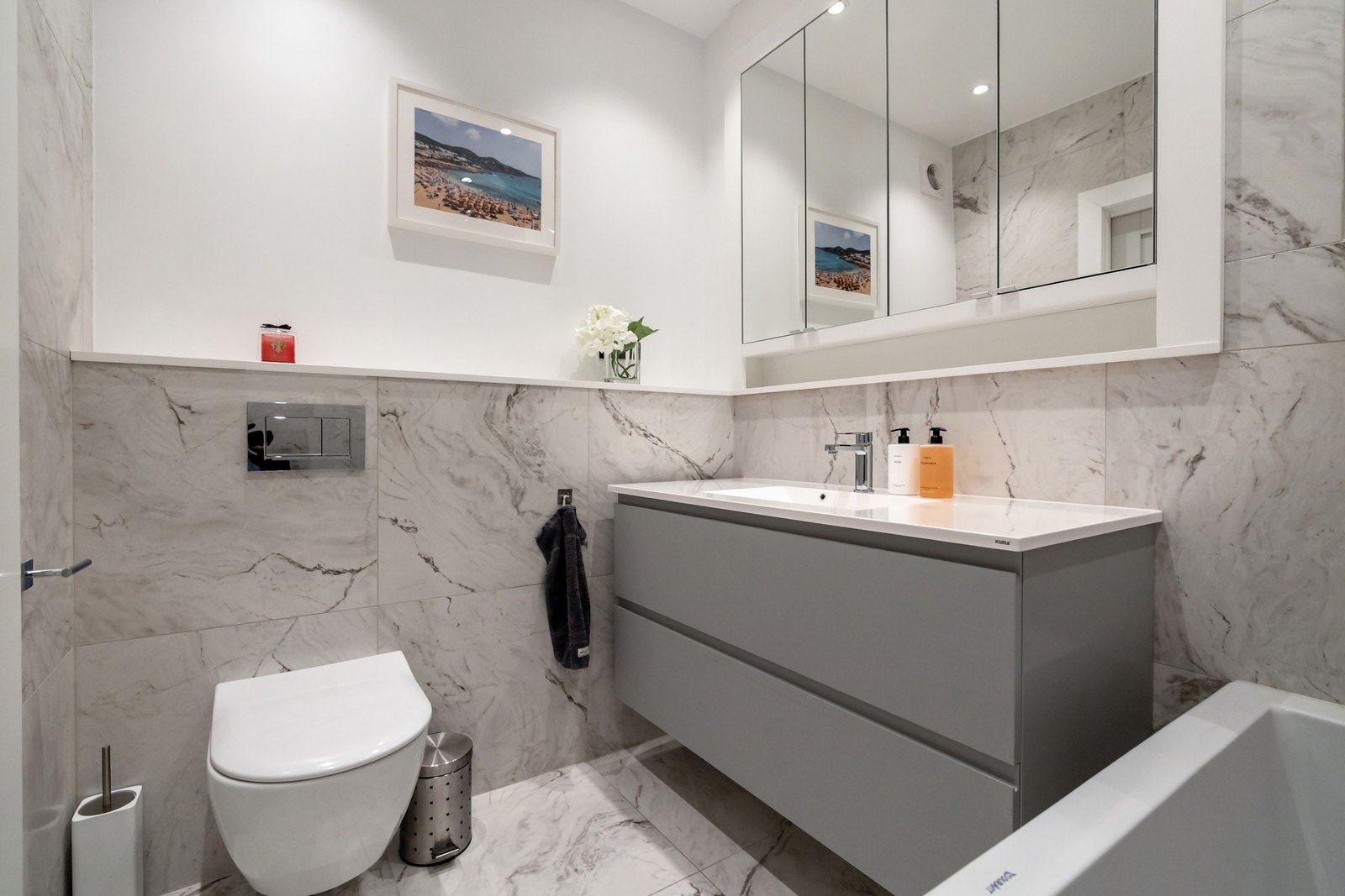 Badet er flislagt med store fliser på gulv og vegger, og er innredet med vegghengt toalett, servantinnredning med oppbevaring. Galleribilde