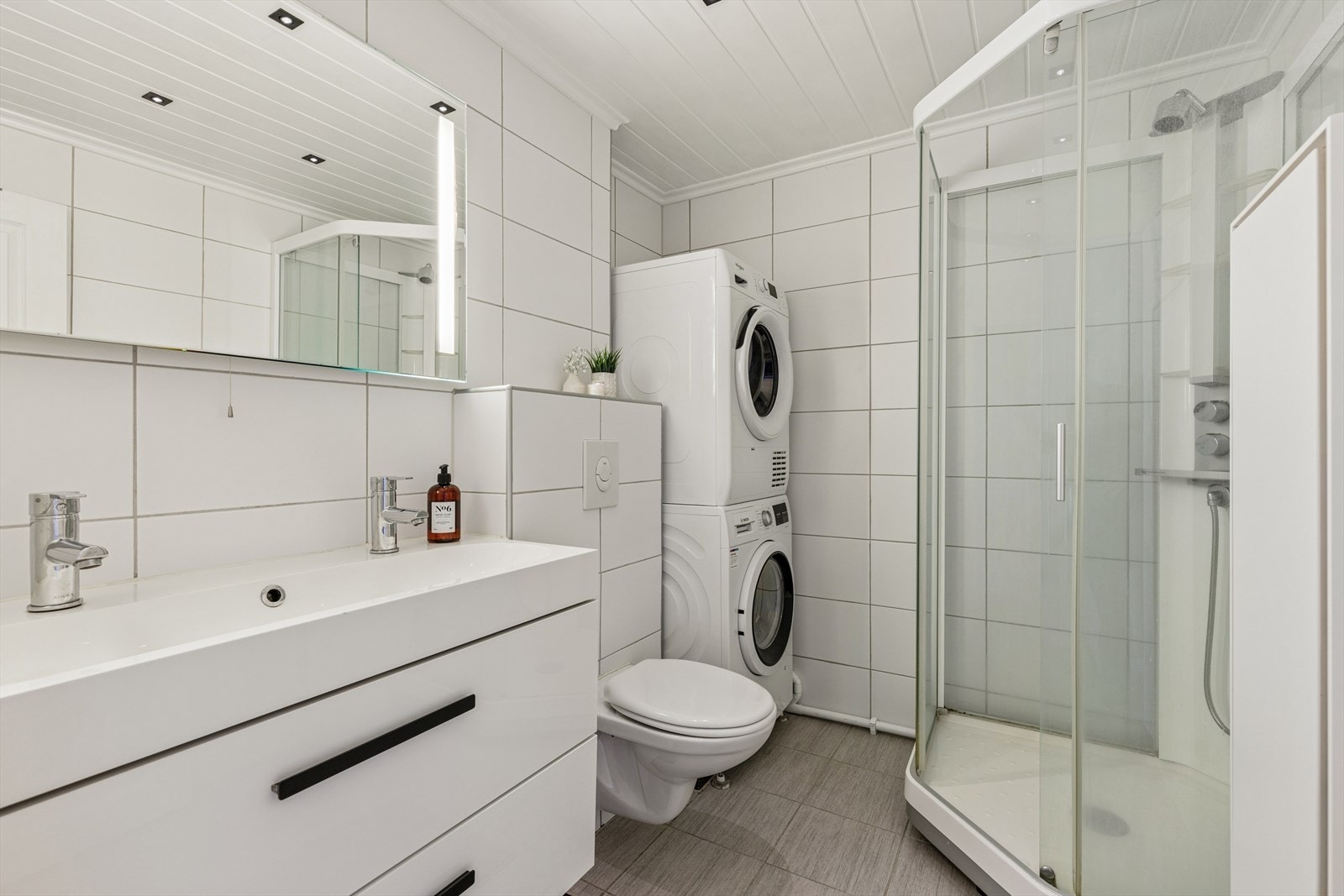 Badet er flislagt, utstyrt med vegghengt toalett, dobbeltservant med underskap, opplegg for vaskemaskin, dusjkabinett og vegghengt skap. Galleribilde