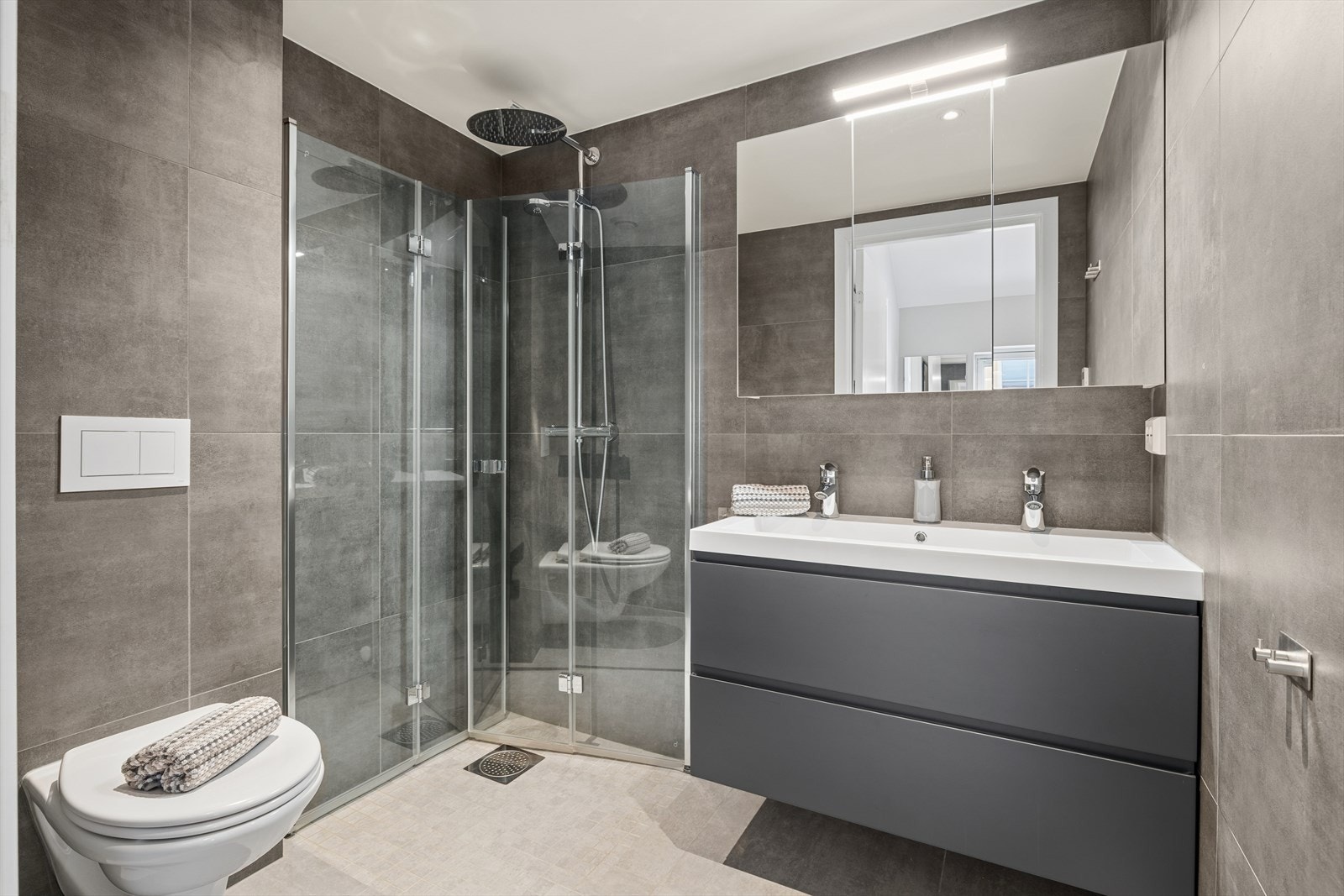 Badet har flislagte overflater, dusjhjørne med glassvegger, vegghengt toalett og vannbåren gulvvarme. Galleribilde