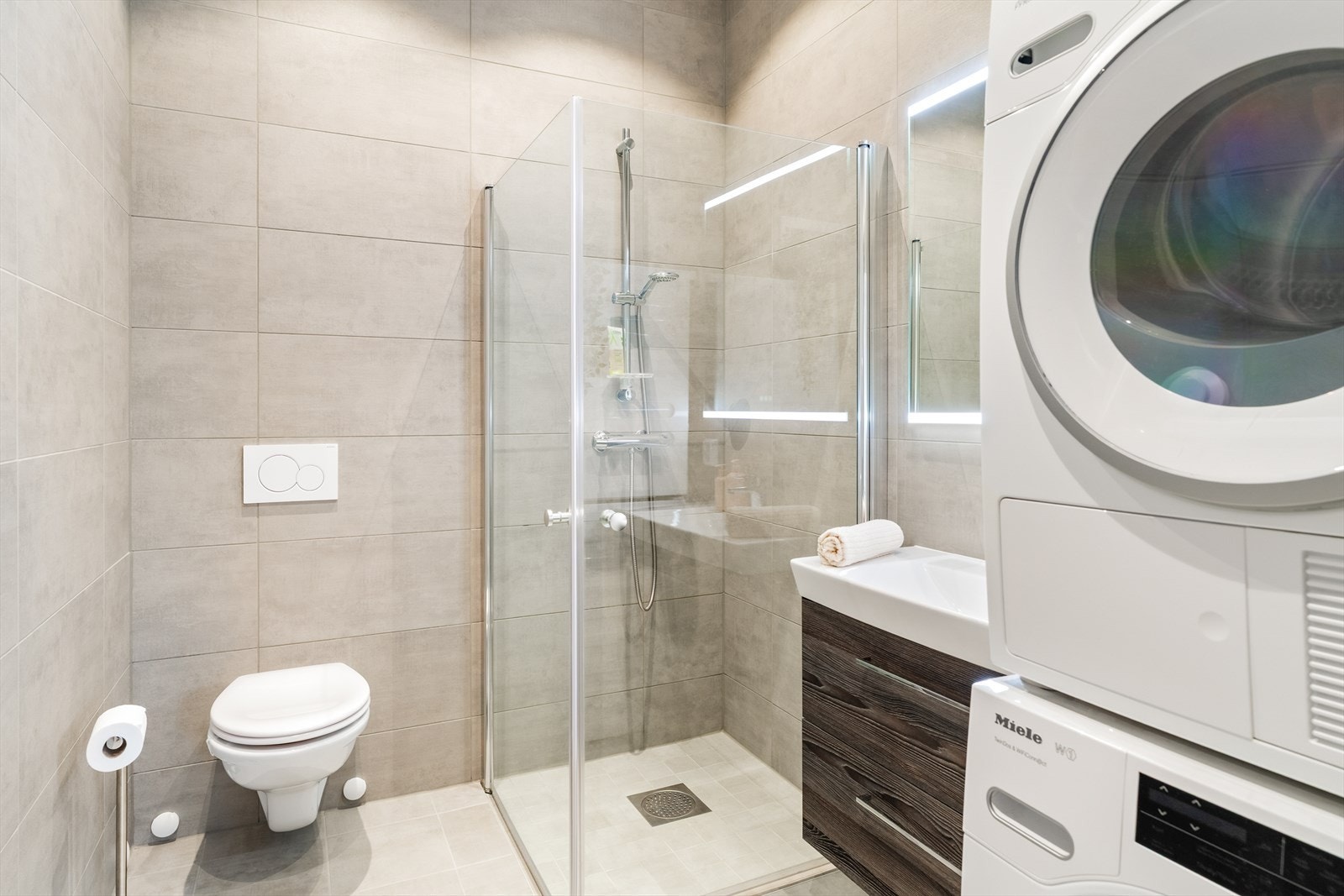 Badet er innredet med servantinnredning, vegghengt toalett, dusjløsning med glassvegger og opplegg for vaskemaskin. Galleribilde
