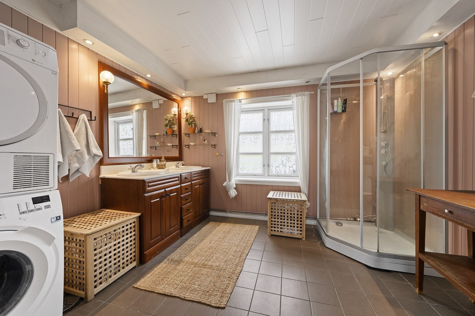 Romslig baderom med flislagt gulv og panel på vegger. Baderommet er utstyrt med dobbel servant over innredning, dusjkabinett og vegghengt toalett. På badet er det opplegg til vaskemaskin og tørketrommel Galleribilde