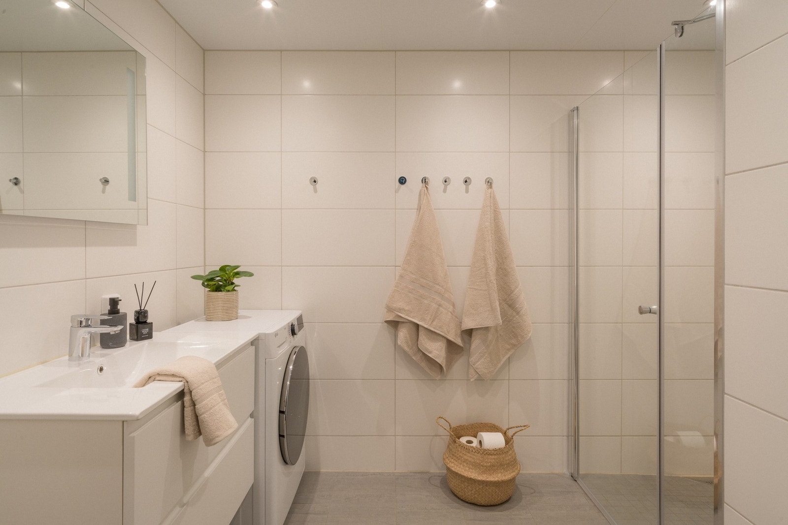 Badet har en delikat utforming og består av en heldekkende servant på hvit skuffeseksjon, speil med integrert belysning, et veggmontert toalett, og en dusjnisje med glassdør. Galleribilde