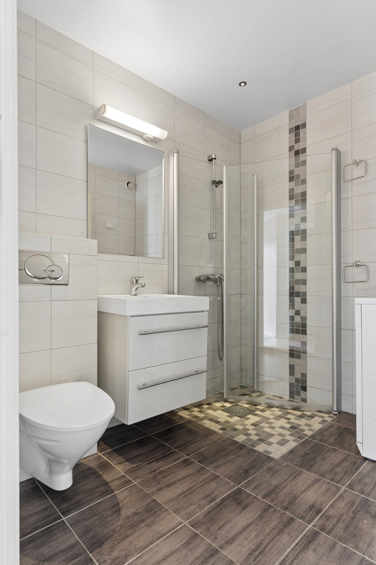 Leiligheten byr på et delikat flislagt bad med en praktisk og funksjonell utforming. Badet er innredet med vegghengt toalett, servant med underskap og speil, samt dusjløsning med glassvegger. Galleribilde