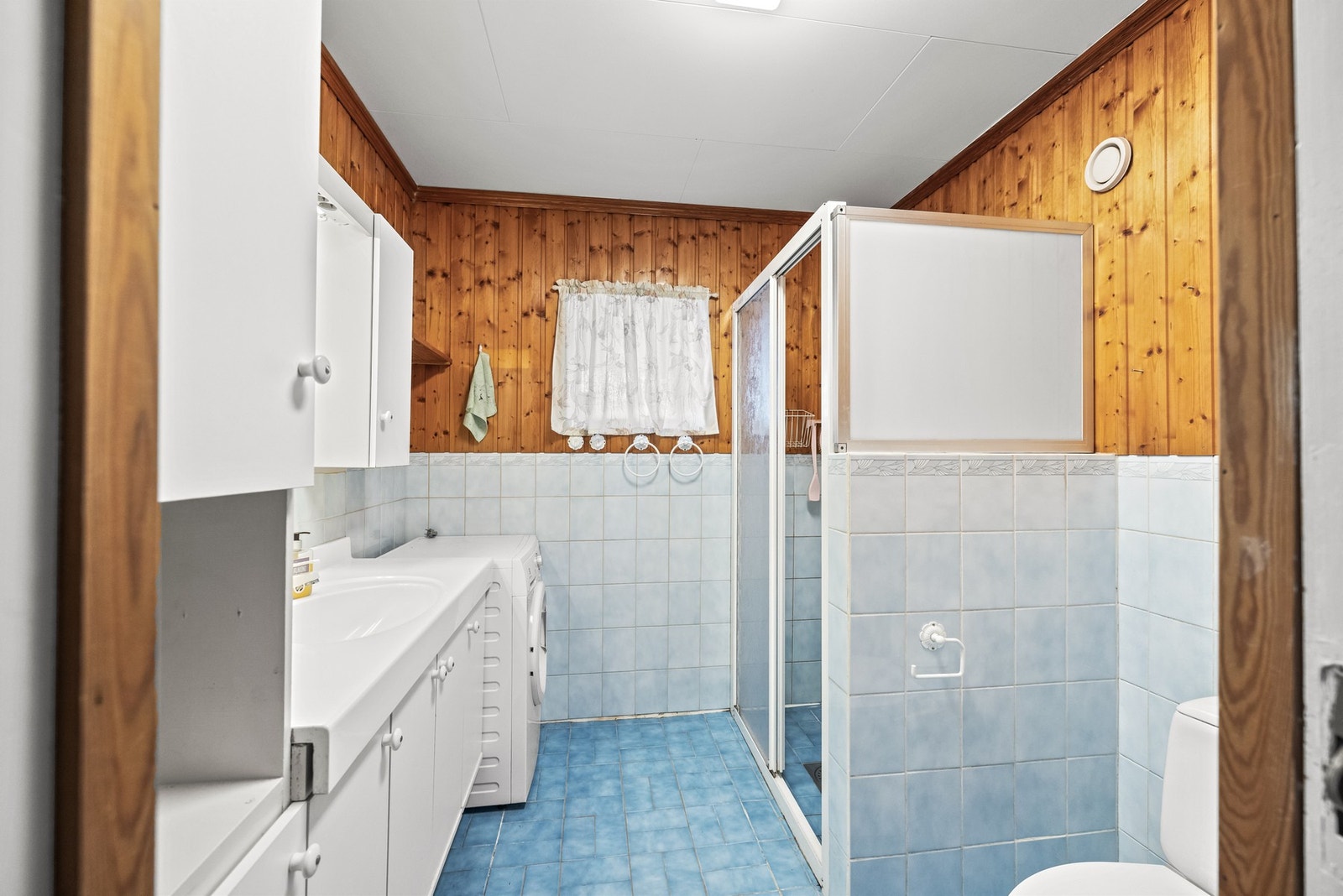 Bad i hoveddel med flislagte overflater, servantinnredning, dusjhjørne og opplegg for vaskemaskin. Galleribilde