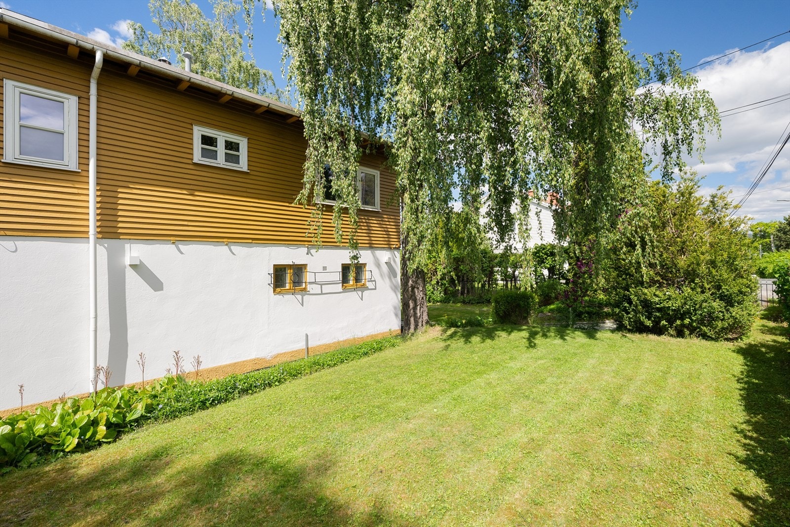 Fasaden er kledd med en kombinasjon av trepanel og pusset mur, som gir huset sitt karakteristiske uttrykk. Flat plen i forhagen med morgensol Galleribilde