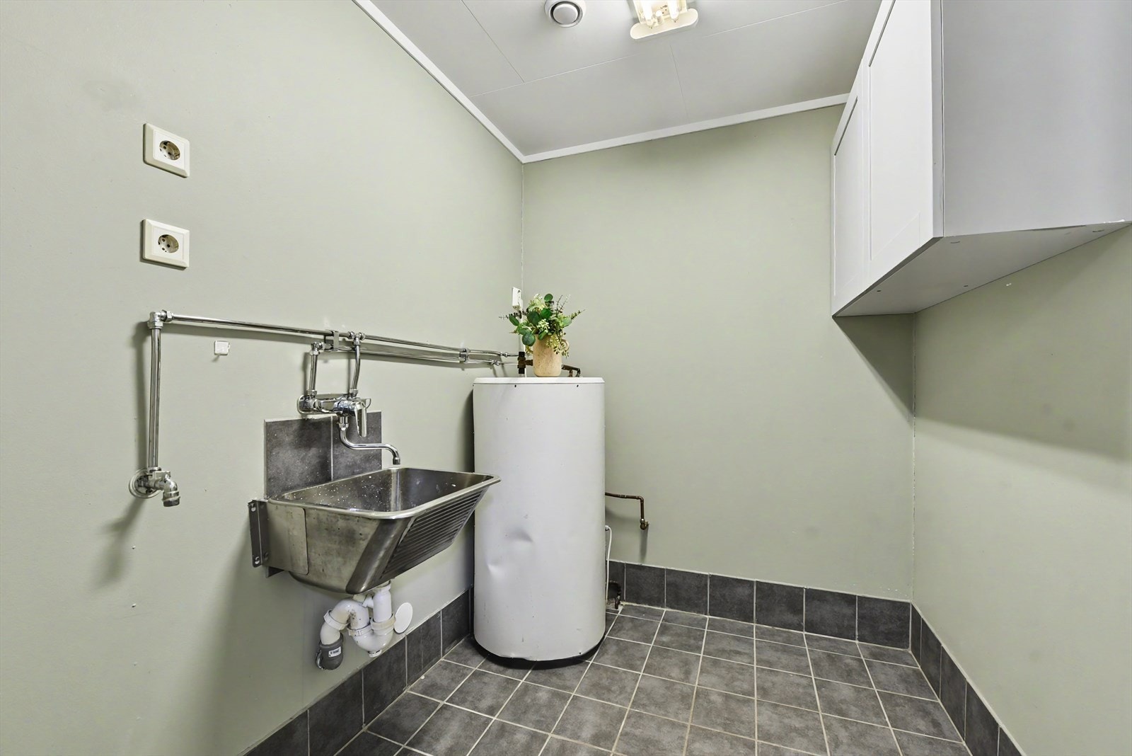 Et separat vaskerom med flislagt gulv, utslagsvask og opplegg for vaskemaskin. Galleribilde