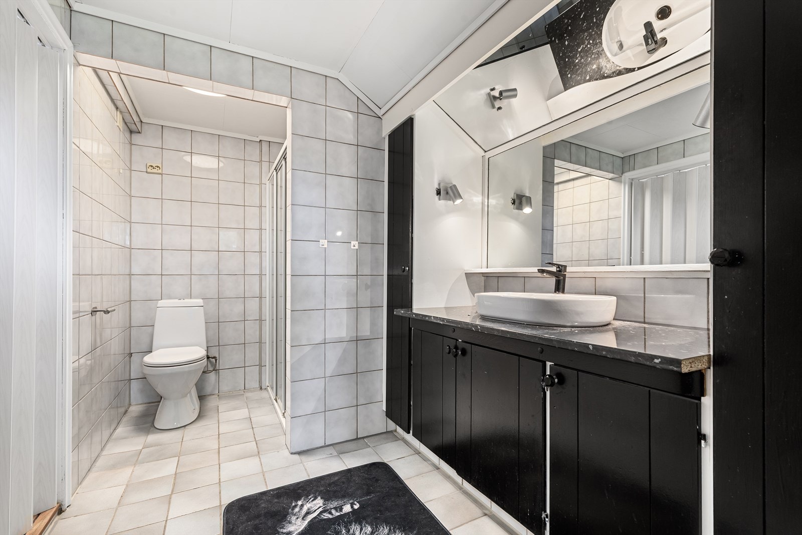 Badetr har innredning med fliser på gulv og vegger. Innredningen har servant med skap under og og ved siden av. Speil og lys. Toalett og dusjhjørne/vegger. Galleribilde