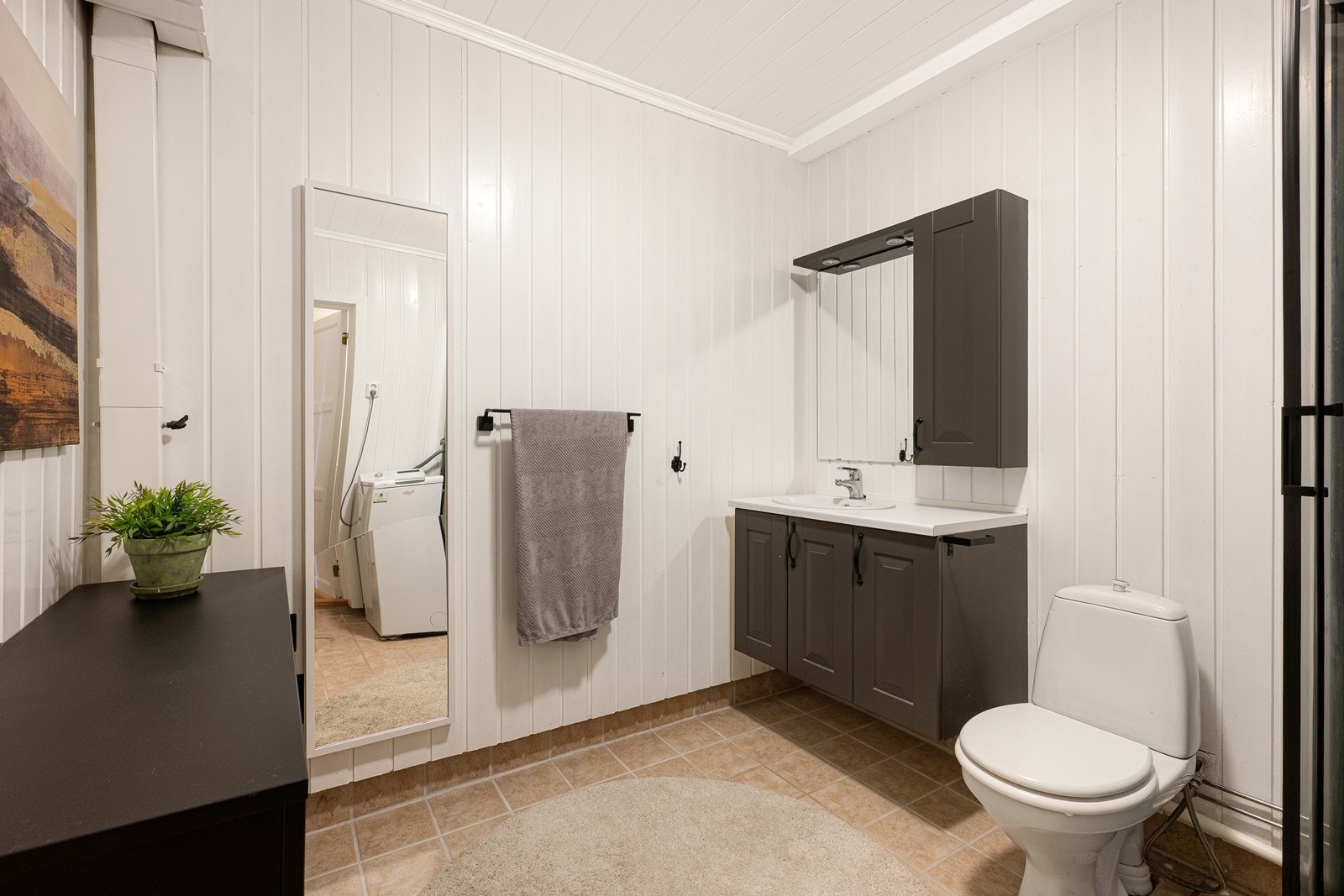 Badet har lysmalt panel på veggene og sandfargede gulvfliser med lun varme i underkant. Innredningen har sorte, profilerte fronter med heldekkende servant over skapseksjon, matchende veggskap og speil med lys i overkant. Galleribilde