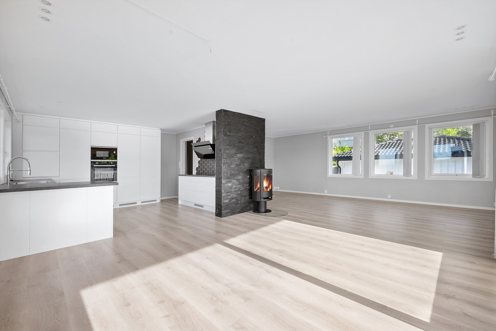 Stuen har åpen løsning mot kjøkkenet, store vindusflater som slipper rikelig med naturlig lys inn og en moderne peisovn som står sentralt plassert i midten av rommet Galleribilde