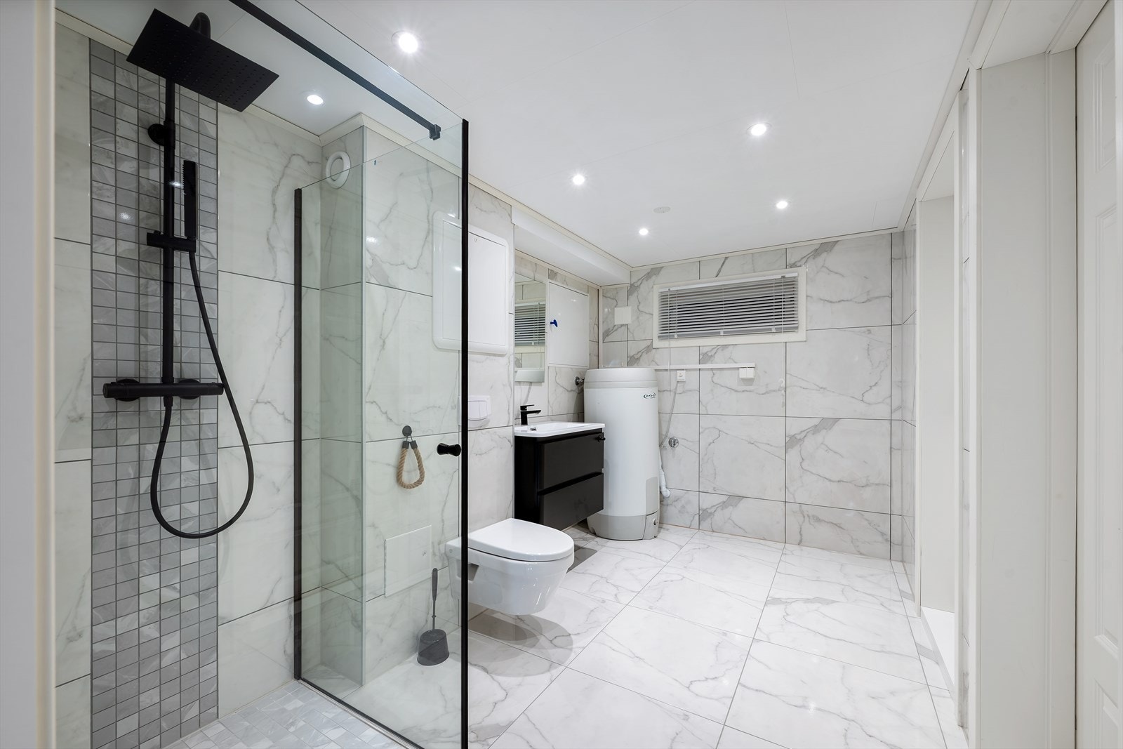 Det kombinerte bad/vaskerommet i kjelleren er pent innredet med fliser på gulv og vegger med underliggende varme i gulv. Badet er videre utstyrt med heldekkende servant, speil, vegghengt toalett, regnfallsdusj og opplegg for vaskemaskin/tørketrommel Galleribilde