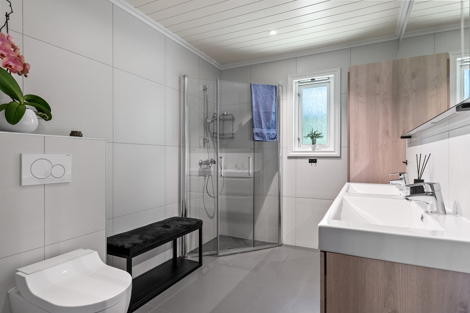 Videre er badet utstyrt med et dusjhjørne med glassdører og vegghengt toalett. Gjennomtenkte løsninger, gode oppbevaringsmuligheter og et tidsriktig design gjør dette til et innbydende bad med høy brukskomfort. Galleribilde