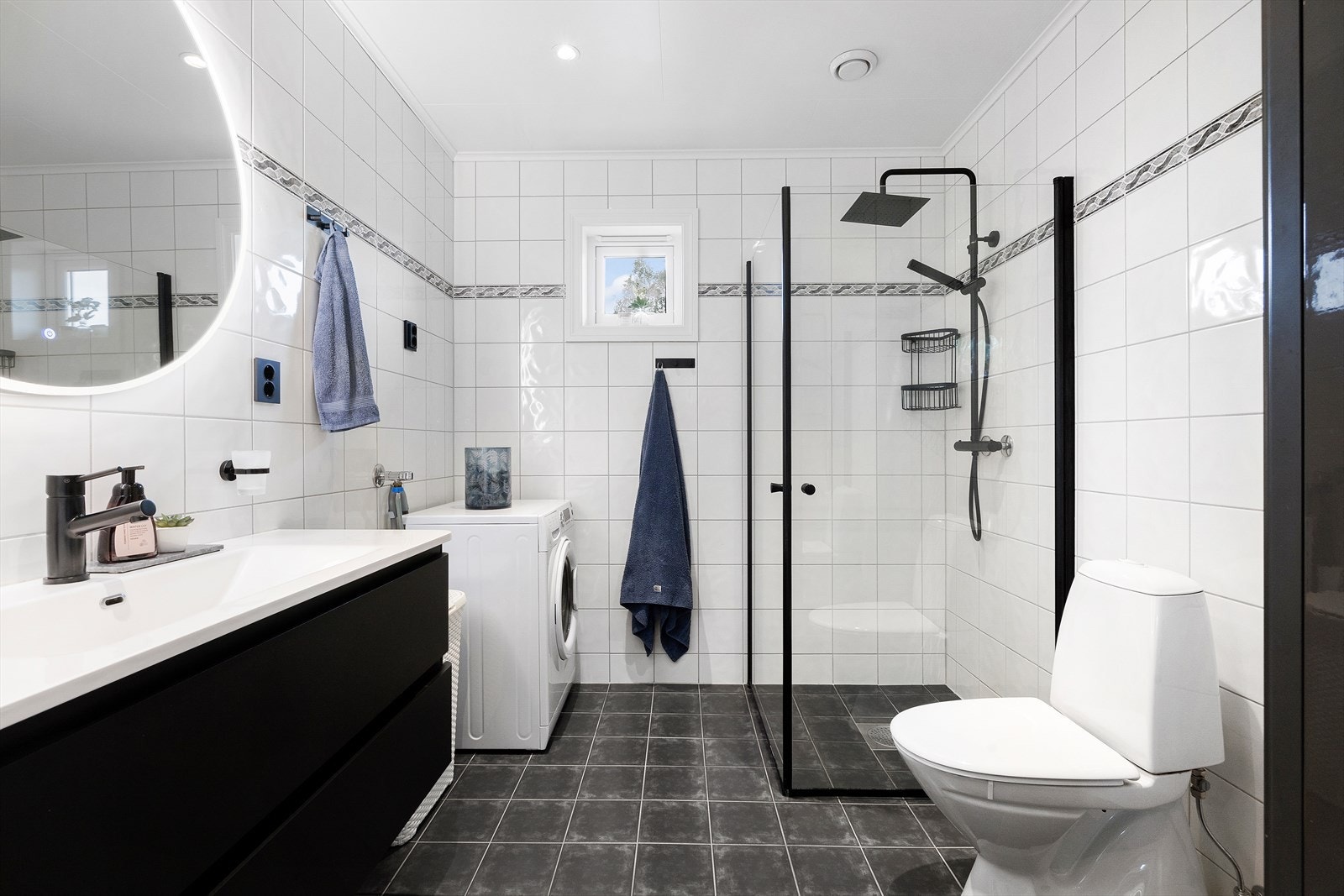 Pent flislagt bad med varme i gulv. Vindu gir sørger for lys og luft. Badet inneholder dusjhjørne, toalett, servant med skuffeseksjon og opplegg for vaskemaskin. Galleribilde