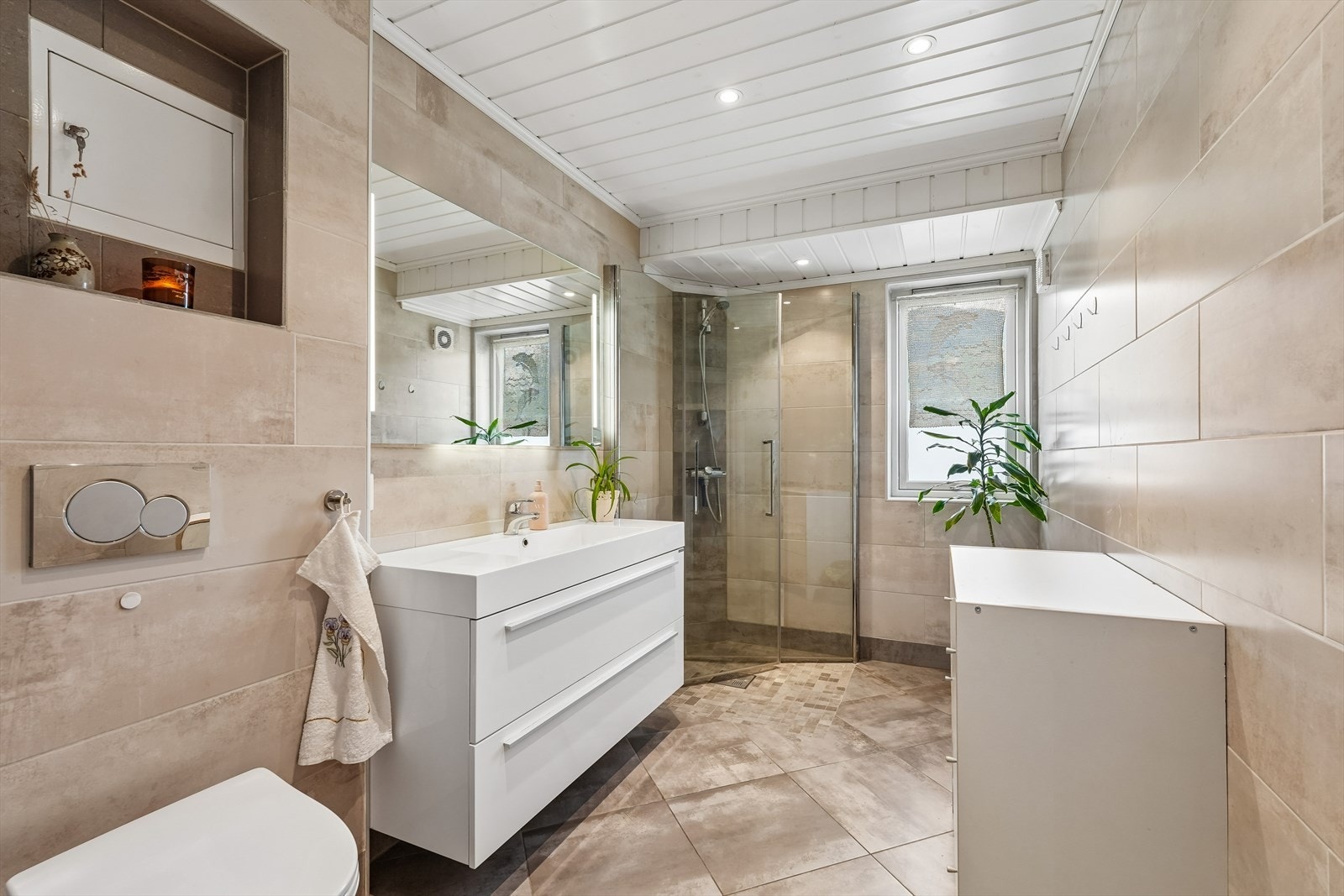 Badet holder en moderne standard med helflisede overflater og varmekabler i gulv. Badet er utstyrt med vegghengt toalett, innredning med helstøpt servantplate, speil og dusjsone. Galleribilde